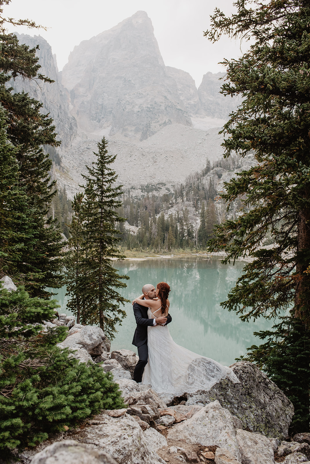 wedding day picture of a bride and groom embracing each other while standing on rocks in a forrest and looking out into a crystal cler lake with the Grand Tetons towering over them