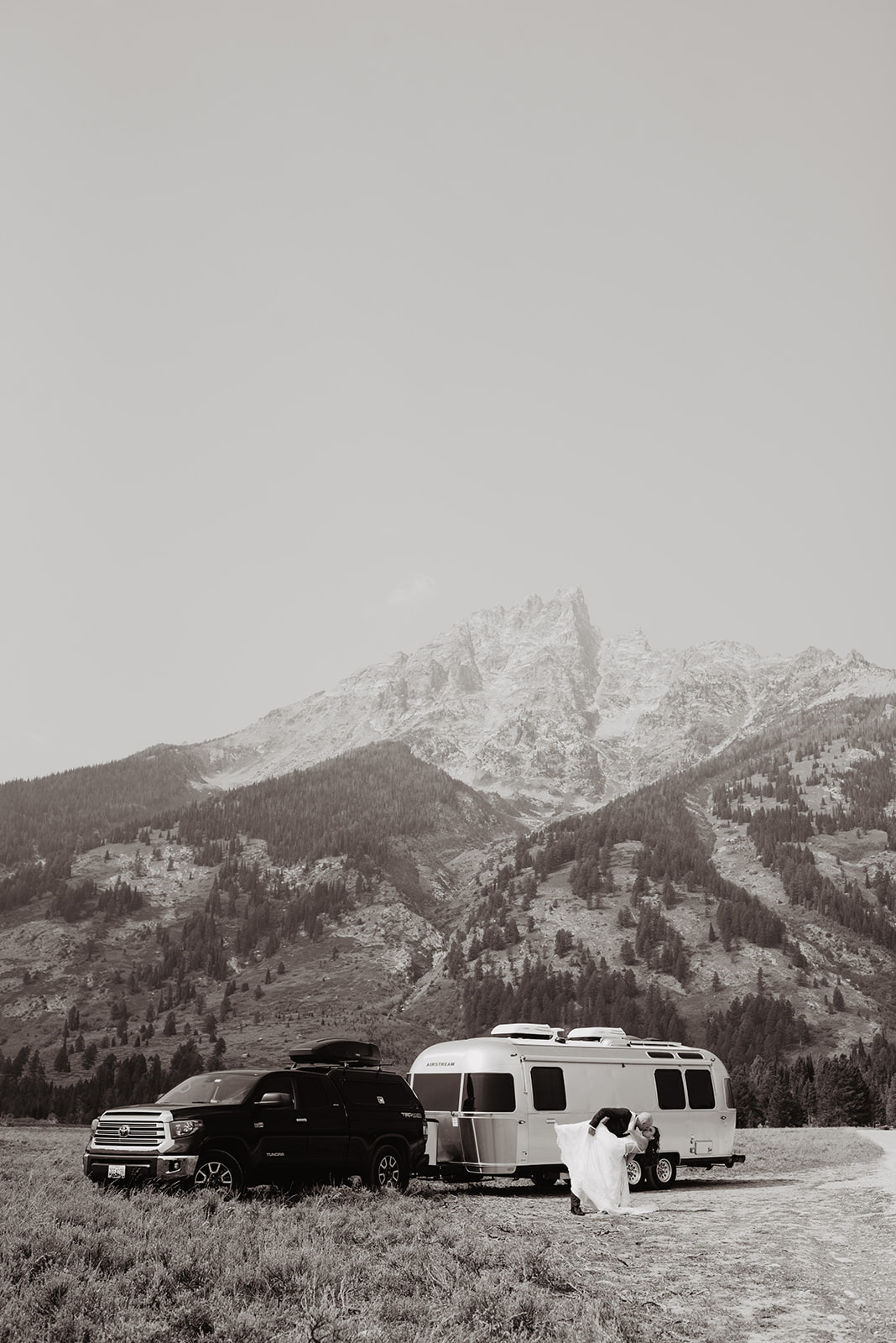 black and white image of a truck and airstream in a field with the Grand Tetons behind with groom dipping his bride and kissing her in front of the airstream