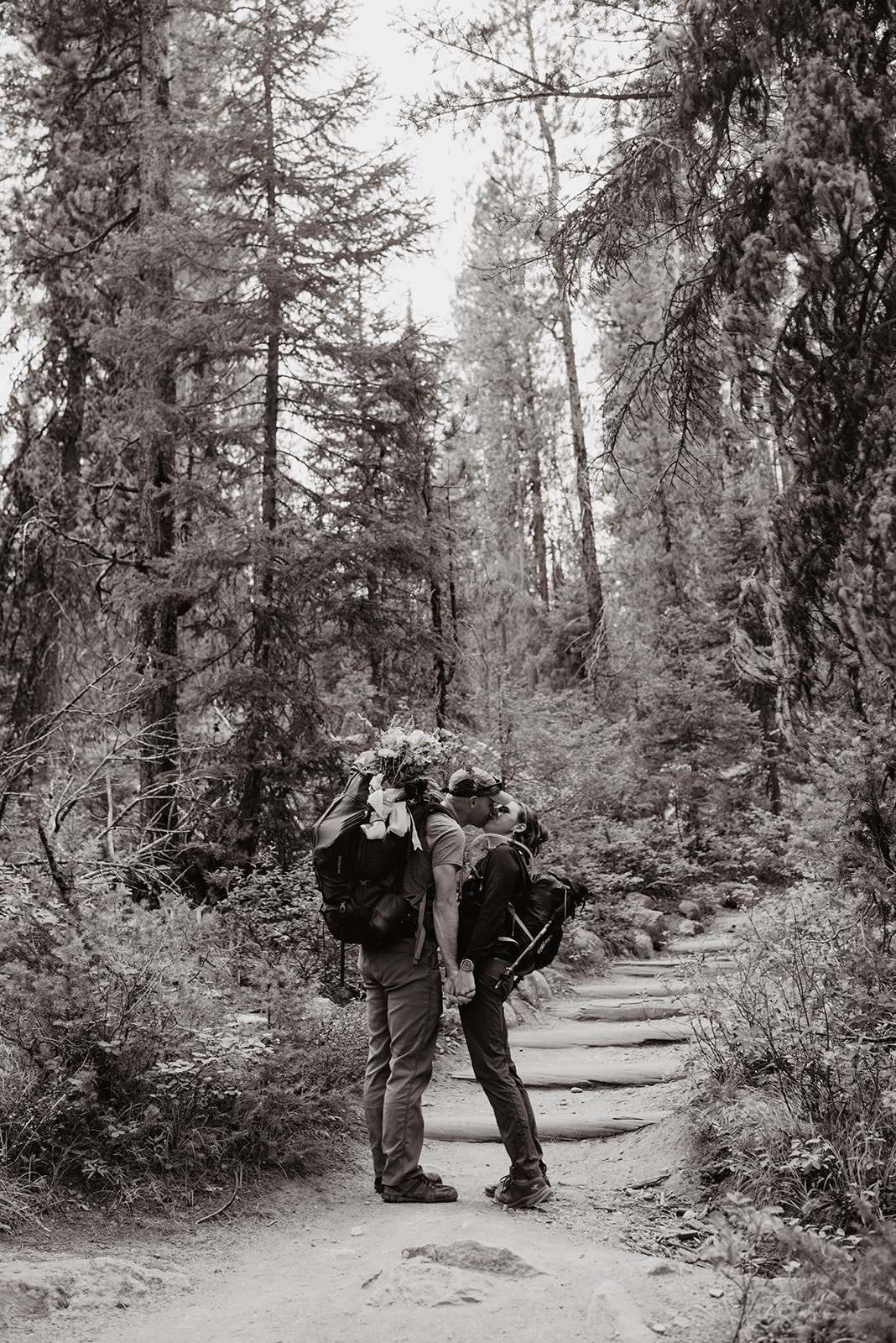 black and white wedding photo of bride and groom kissing each other on a dirt trail in the Grand Tetons with tall native trees surrounding them