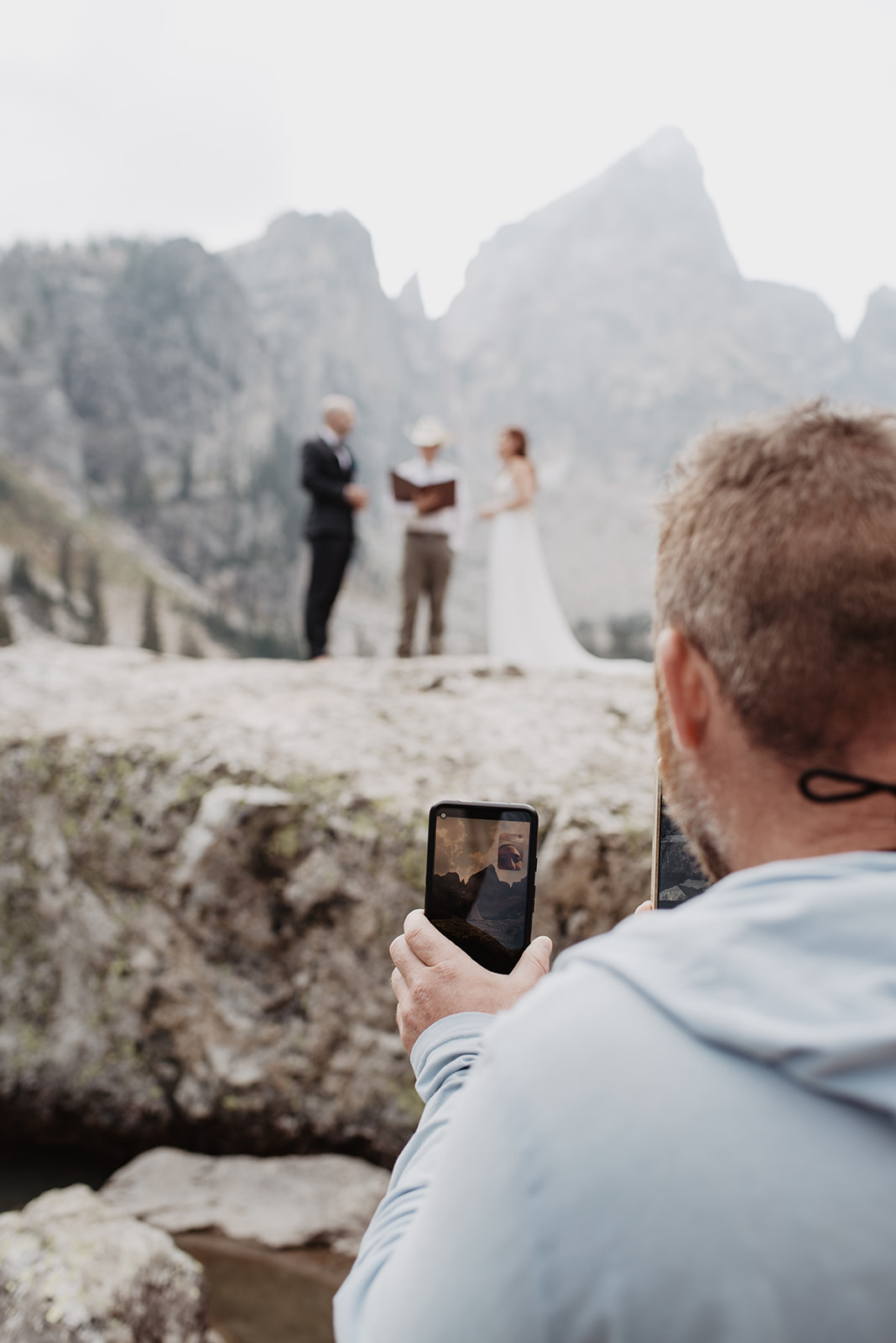adventure wedding day in the Grand Tetons with the bride and groom standing with their officiant on a large rock with the Teton mountain behind them as a man holds their iphone to facetime with a friend