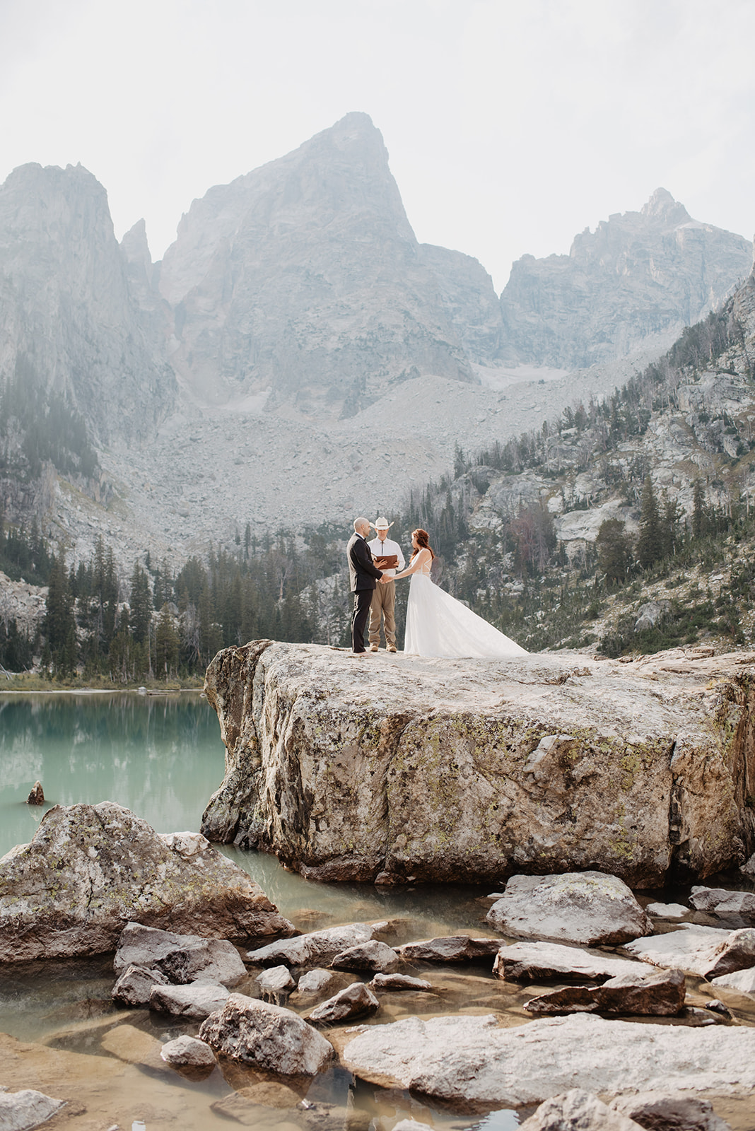 elopement ceremony inthe Grand Tetons in the fall with bride and groom holding hands on a rock with their officiant with the Tetons mountain range right behind them as they say their vows over a glacier lake 