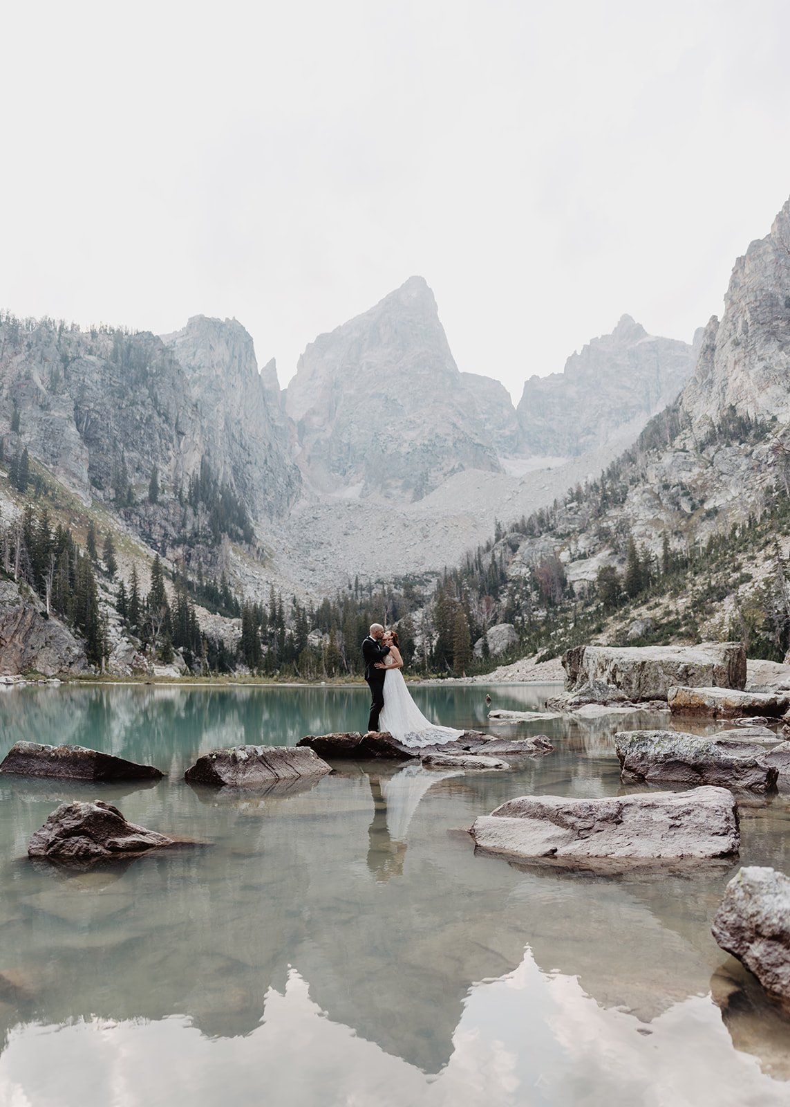 man and woman standing on rocks in a glacier lake in the valley of the Tetons mountains with aqua blue water surrounding them as they kiss on their wedding daywith the Tetons in the background and reflecting into the lake