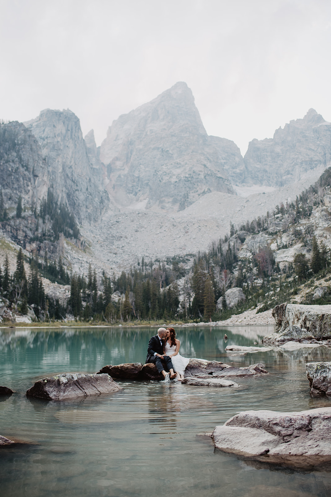 bride and groom sitting on a rock in a aqua blue glacier lake in the Grand Teton National Park kissing each other with the Tetons towering over them in the background