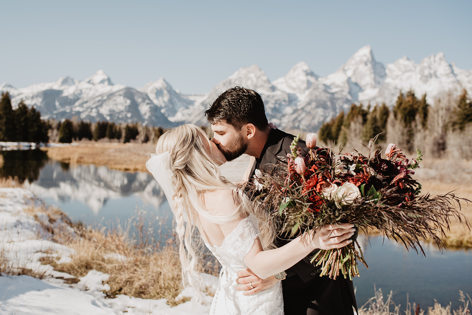 winter wedding in Jackson Hole Wyoming with bride and groom kissing each other next to a river with the Grand Tetons mountain rnae in the background and the bride holding her red floral wedding bouquet