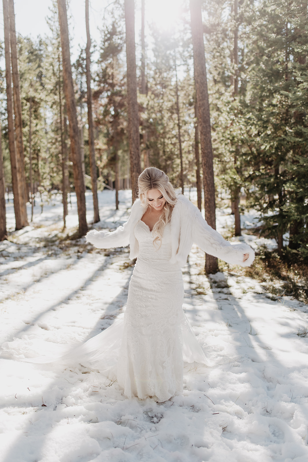 bride walking through the forest in the Tetons on her wedding day in the winter as she wears a lace mermaid gown and a white fur coat with the light coming from behind her and casting the long shadows from the trees over her