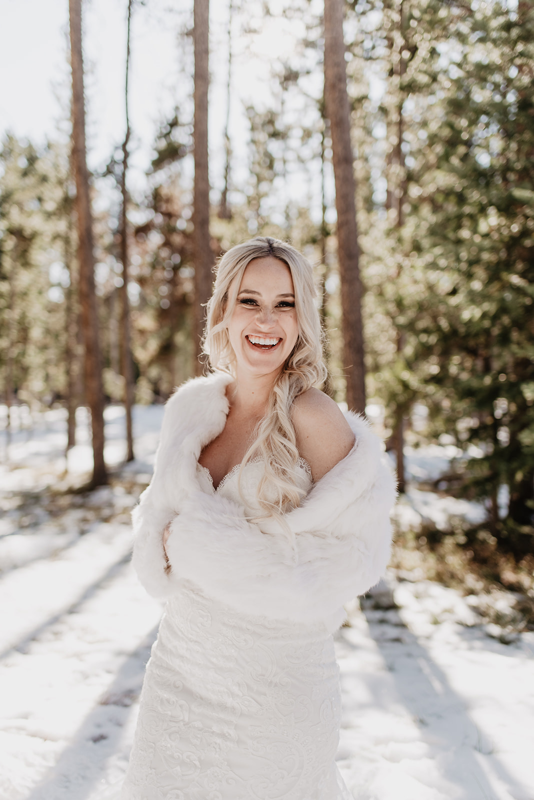 very happy bride in the woods in the winter in Jackson Hole wearing a faux fur coat they drops off her shoulder a bit with her hands to her heart