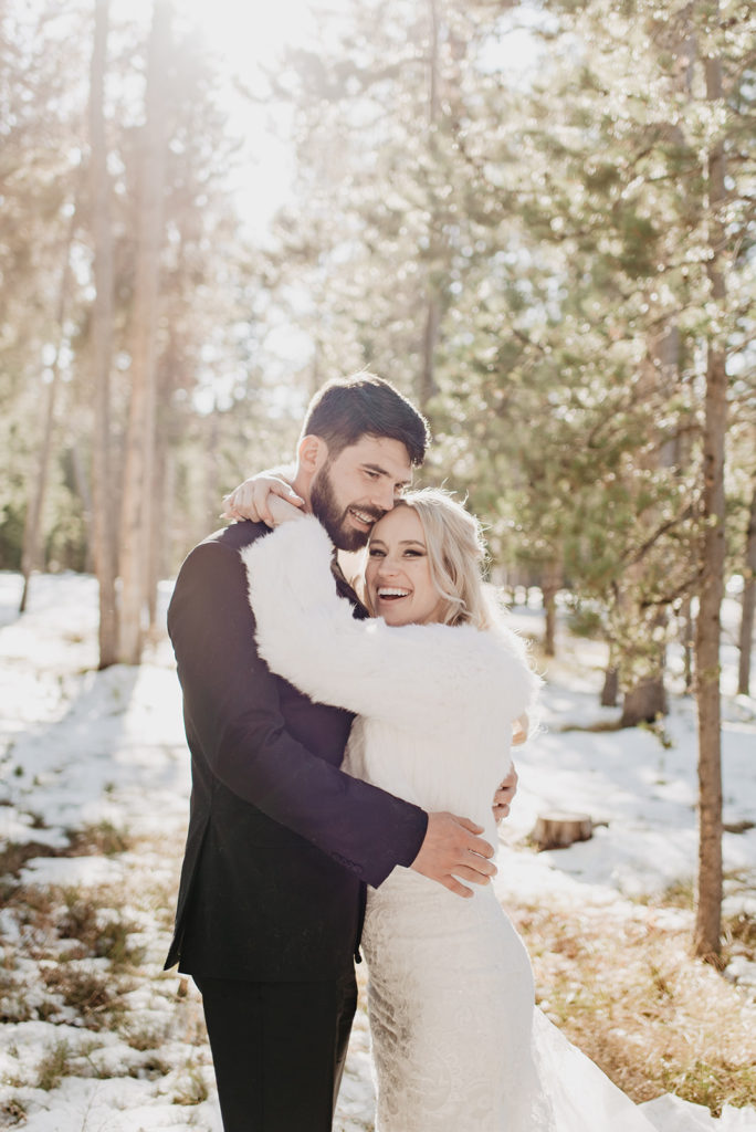 bride in a lace wedding gown and a faux white fur jacket holding her groom around the neck as the groom who is in a black suit holds her waist as they stand in a snowy forest in Jackson Hole with the sun flickering through the trees