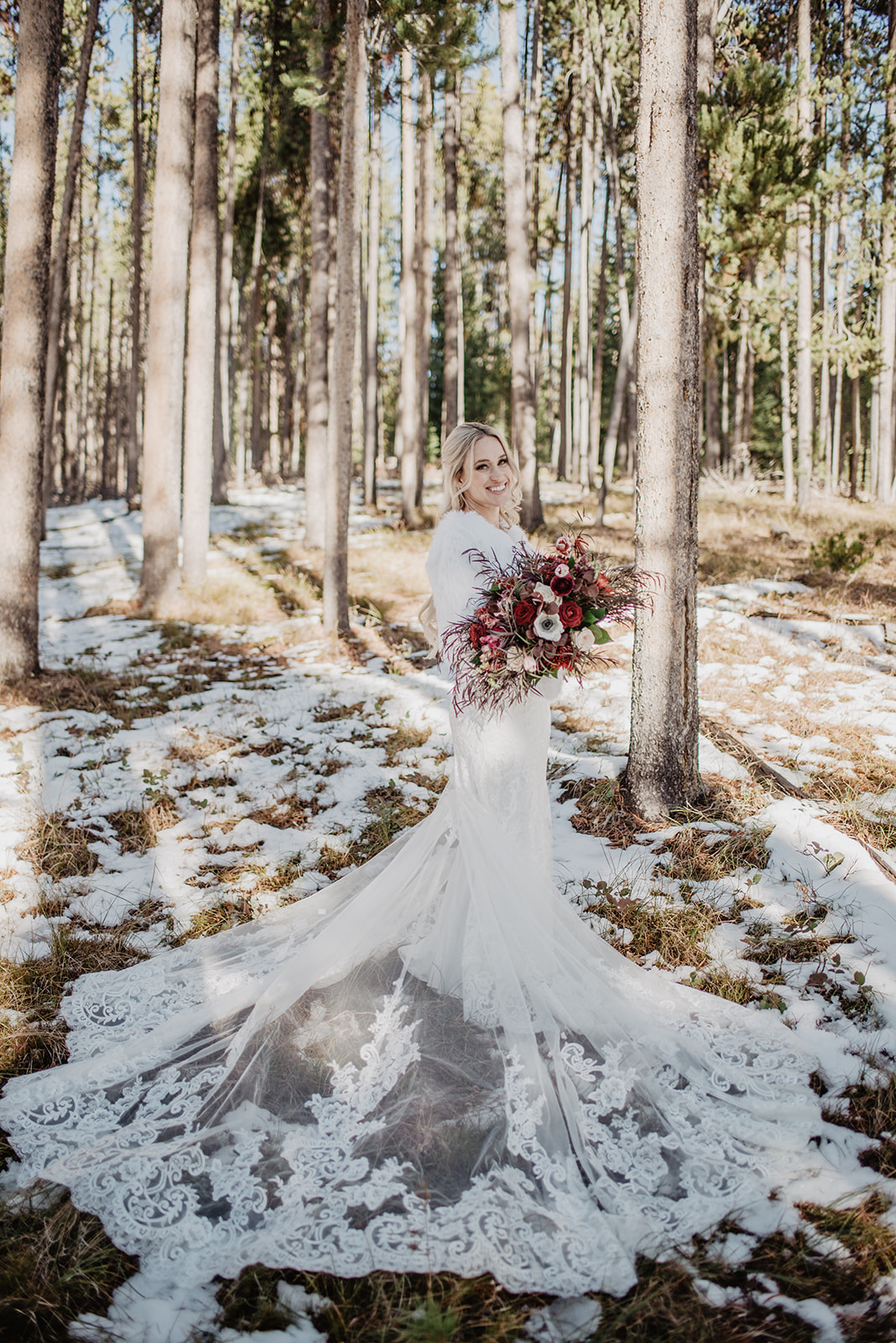 bridal portrait of bride standing in a snowy forest and holding her modern deep red wedding florals as her long lace train is spread out in front of her