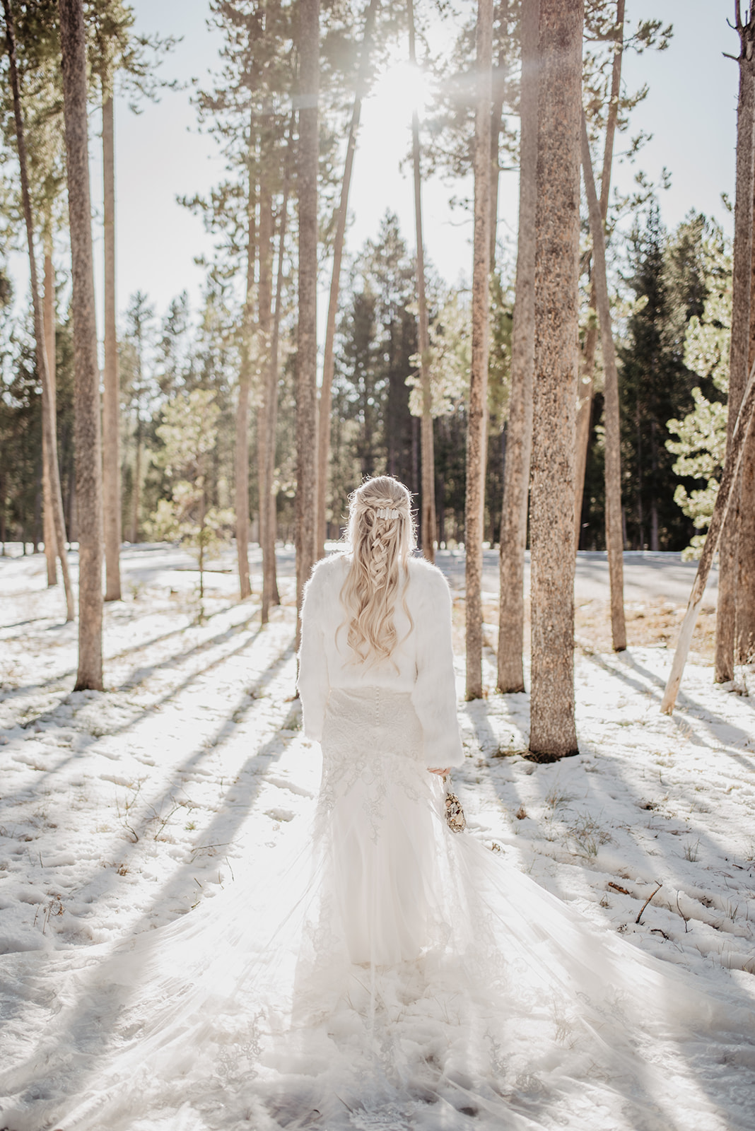 winter bride walking towards a tree line with the sun shinging through to her as she walks away from the camera for her elopement pictures