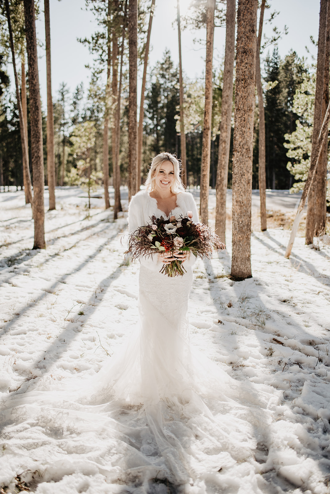 bride standing in the woods with her deep red wedding bouquet as the sun from behind her casts shadows of the trees over her