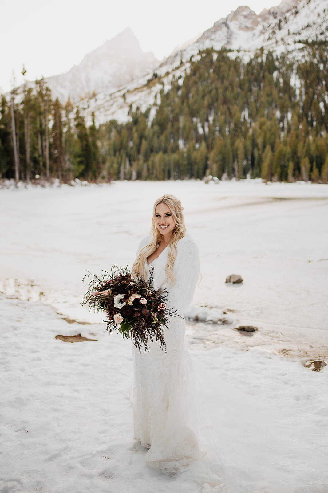 blonde bride in a lace and floral wedding gown and white faux fur coat stands on the edge of a fozen lake with the Grand Stetons behind her with a haze of snowy fog over them, at String Lake in the Winter