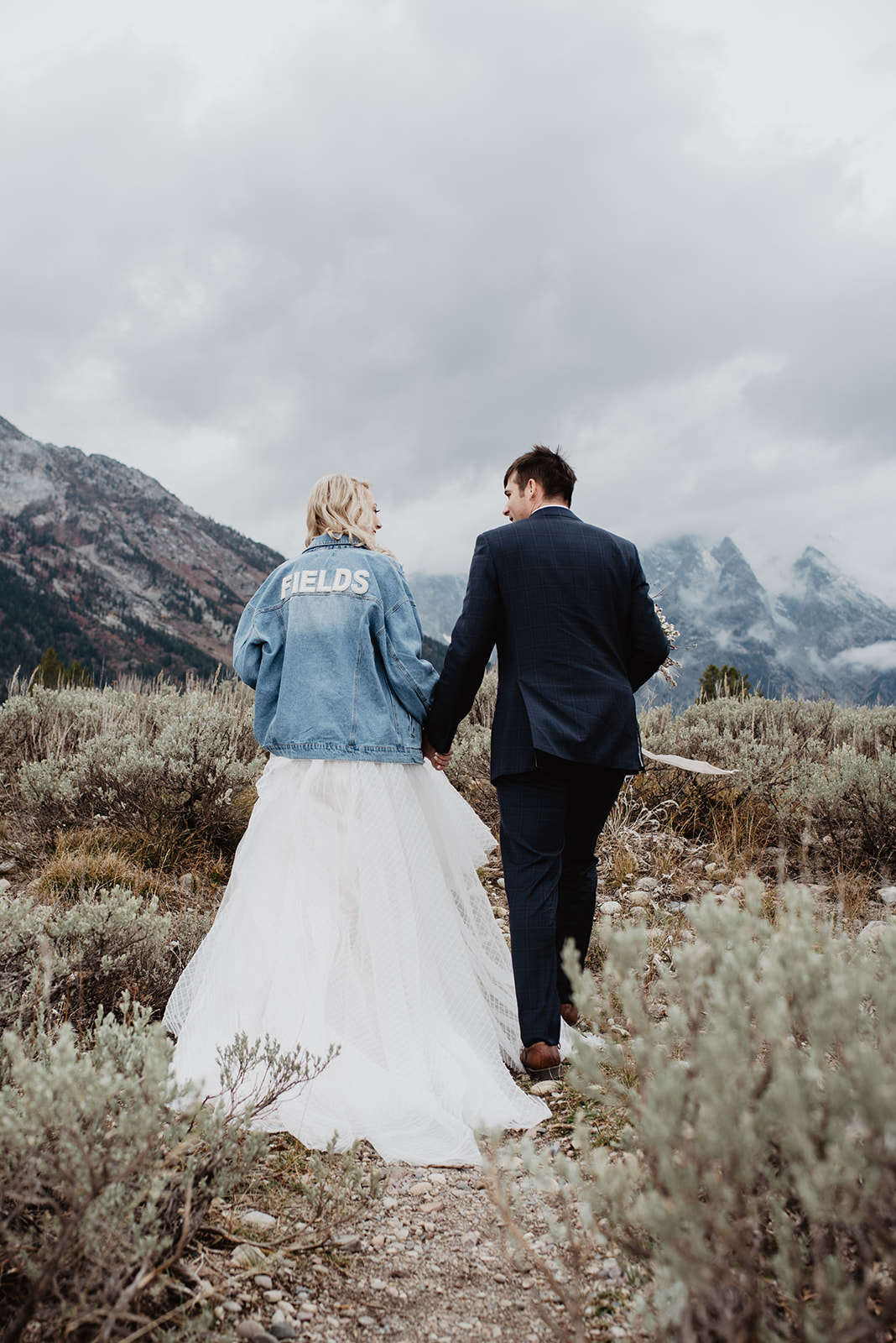 bride in her wedding dress and a denim jacket holding hands with her groom who is in his dark suit as they walk through a field in Teton Valley towards the Grand Tetons mountains