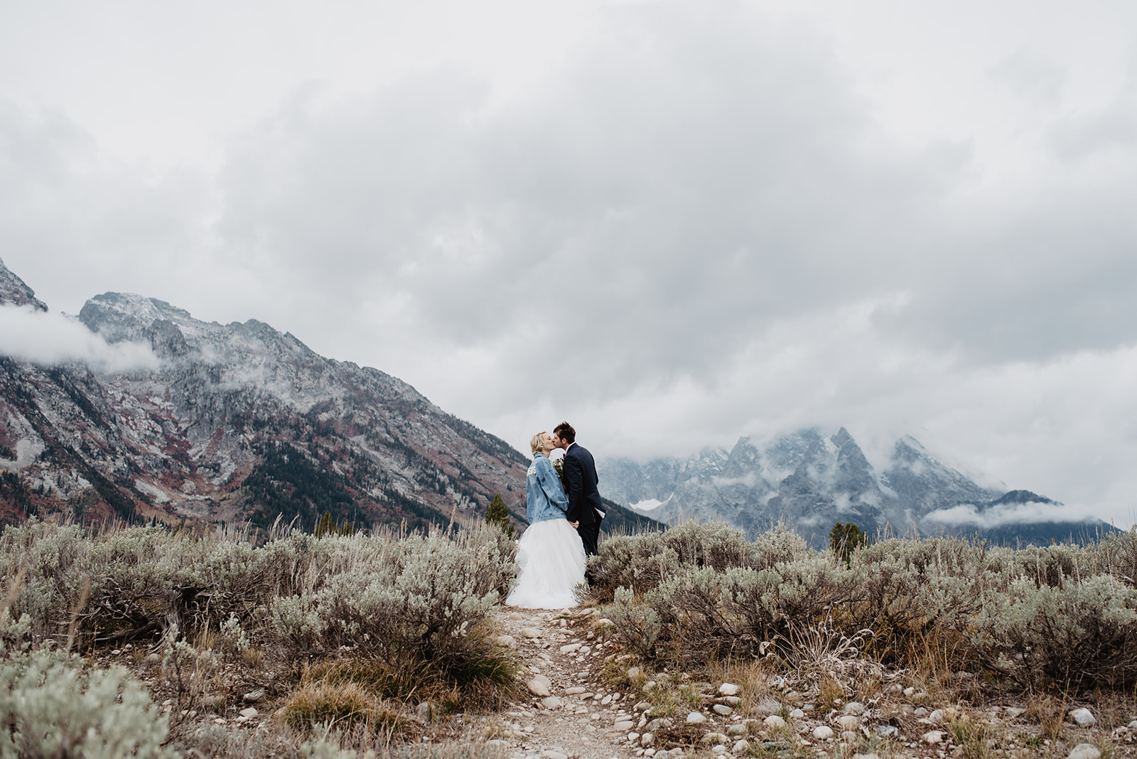 rainy wedding day in the Grand Tetons with bride in a denim jacket kissing her groom as they walk up a hill together. 