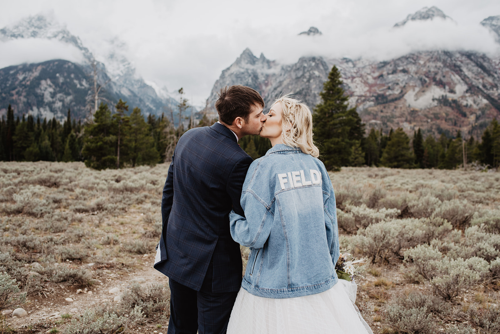 bride kisses her groom in front of the Tetons in Jackson Hole while wearing a jean jacket that says "bride" on the back in white lettering