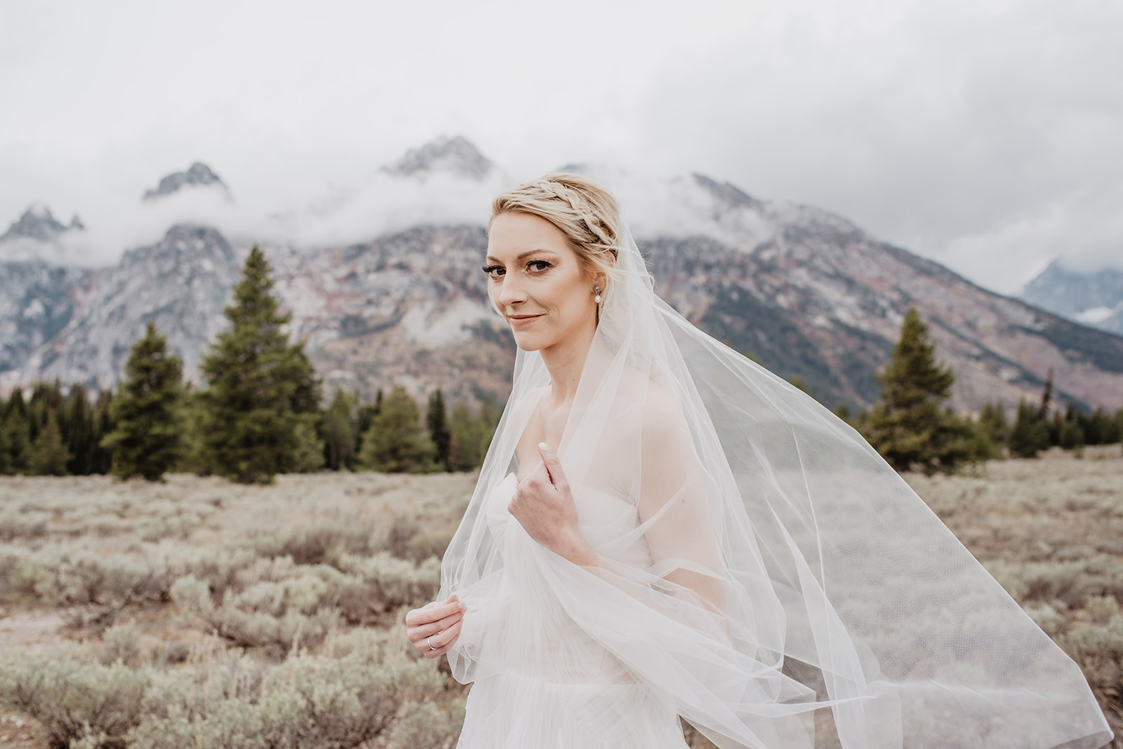 bride standning in fron of the smowy Tetons mountain range smirking at the camera as she holds her veil in place while the wind blows it away from her
