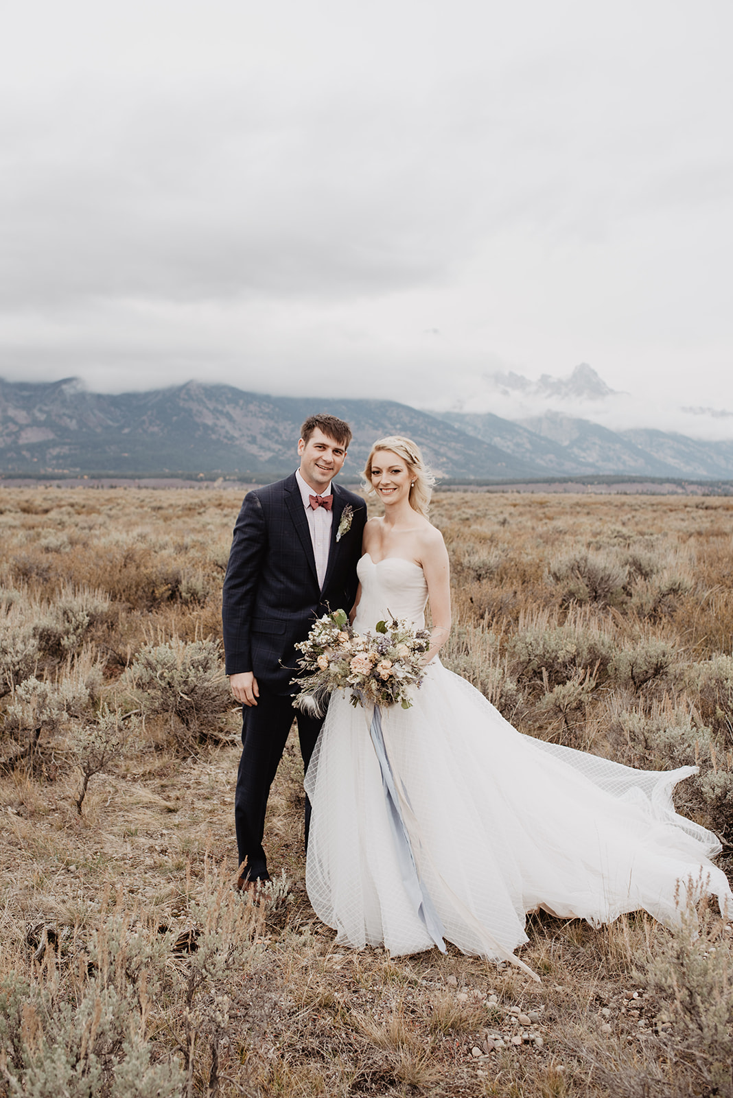 bride and groom standing in a meadow in the Tetons of their wedding day with tie wedding dress and blue suit on smiling at the camera as a fog drifts behind them covering the Tetons mountains