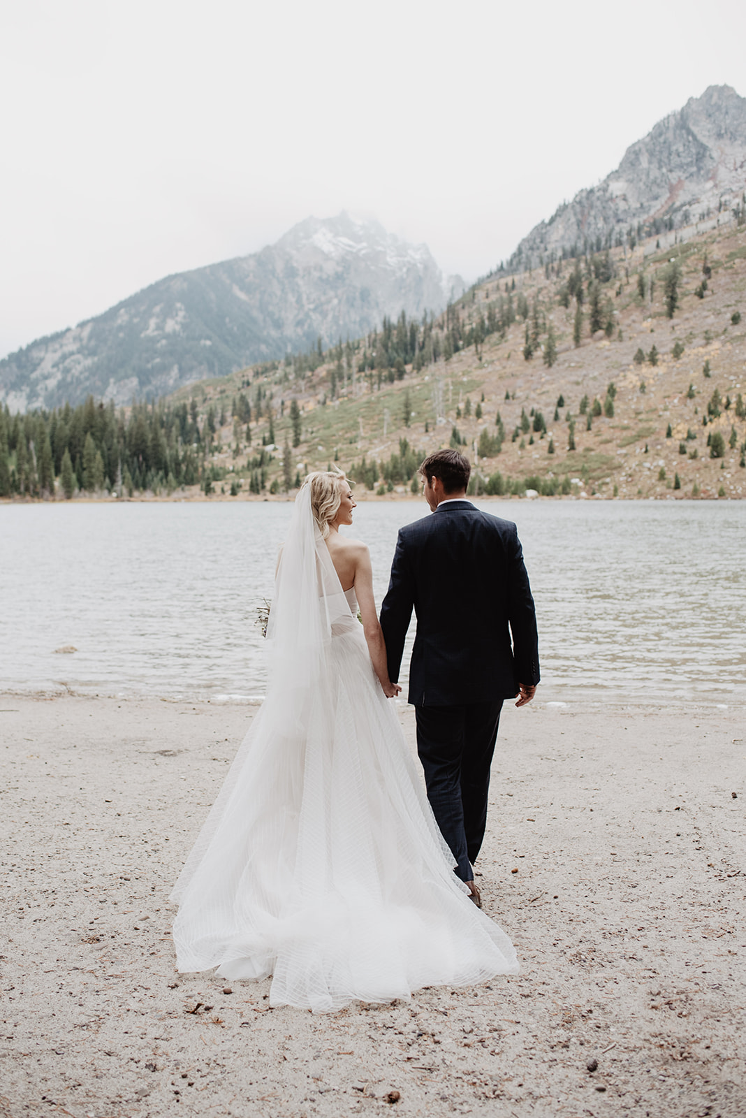 bride and groom in their wedding attire holding hands and looking out to String Lake with the Tetons in the distance with thin fog hovering through the mountains for this Jackson Hole weddings