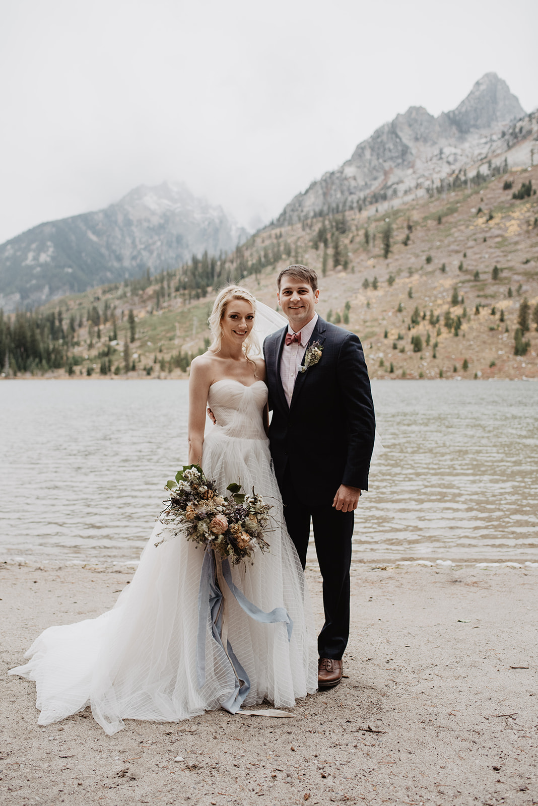 bride and groom standing on the shore of a lake in Jackson Hole with the Tetons behind then as they hold hands and smile for the camera for their adventure session wedding photos