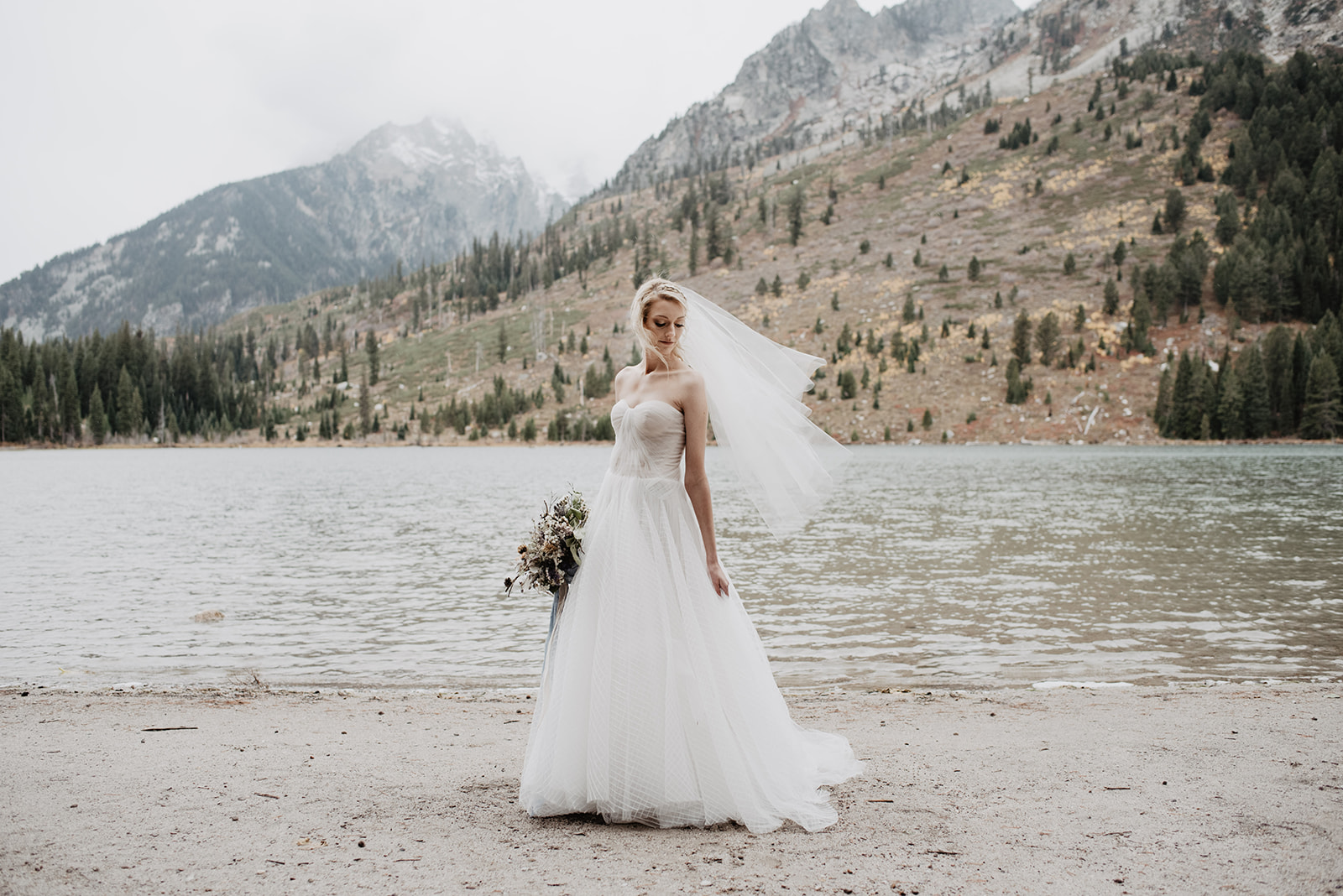 stunning bride at String Lake weddings standing on the shore as the wind blows her gown and veil behind her and she looks off, with mountains behind her and the lake