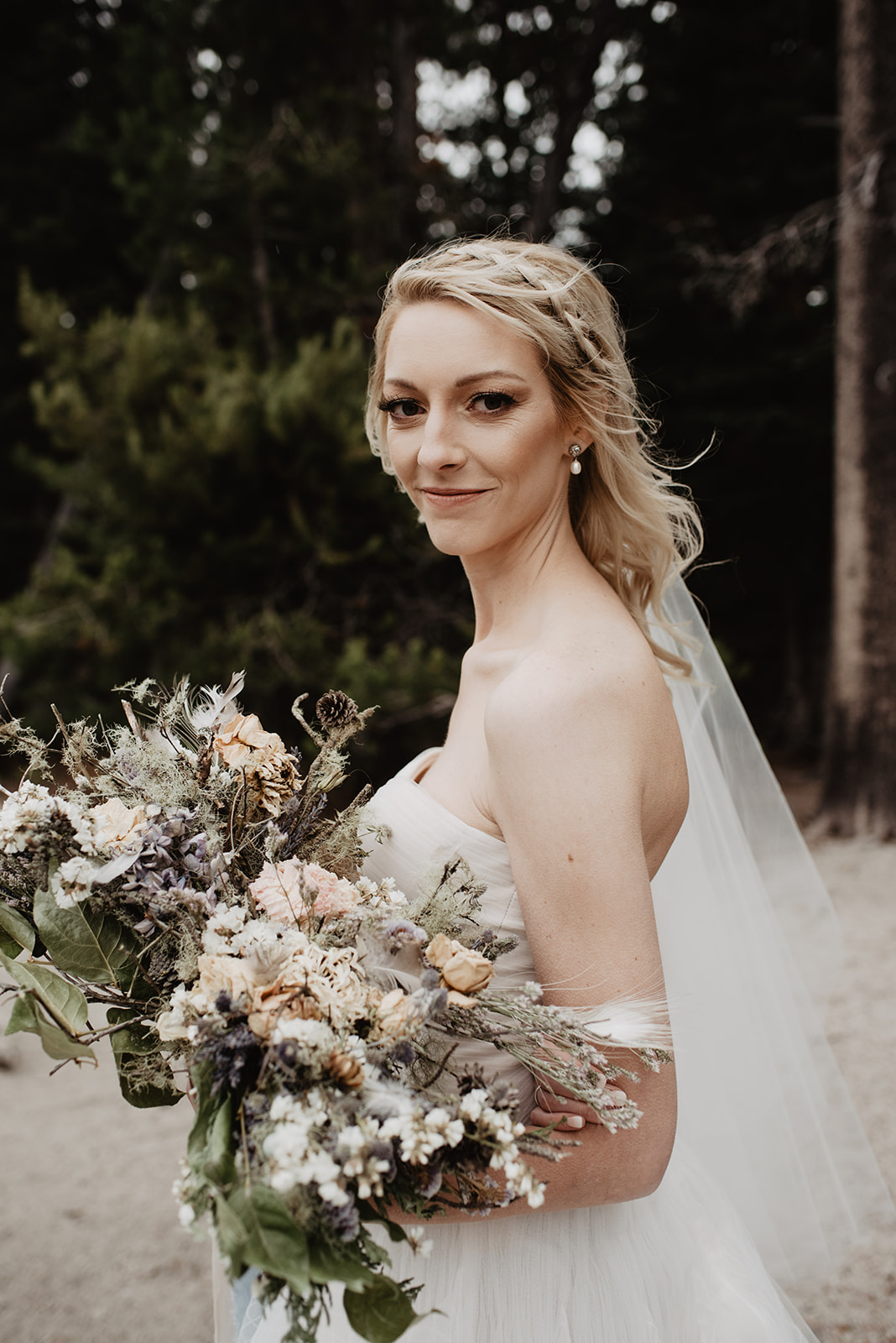 blonde bride looking over her shoulder as she carries her bridal bouquet in Jackson Hole weddings venue, with pine trees towering behind her