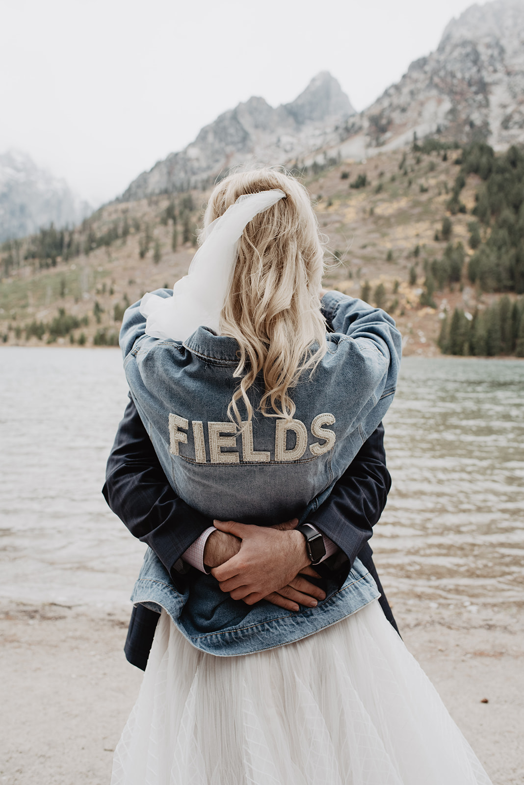 Bentwood Inn wedding venues String Lake wedding photos with bride in a denim jacket with her new last name holding her groom around her shoulder as he holds her around the waist as they stand together in front of the lake for their Jackson Hole weddings
