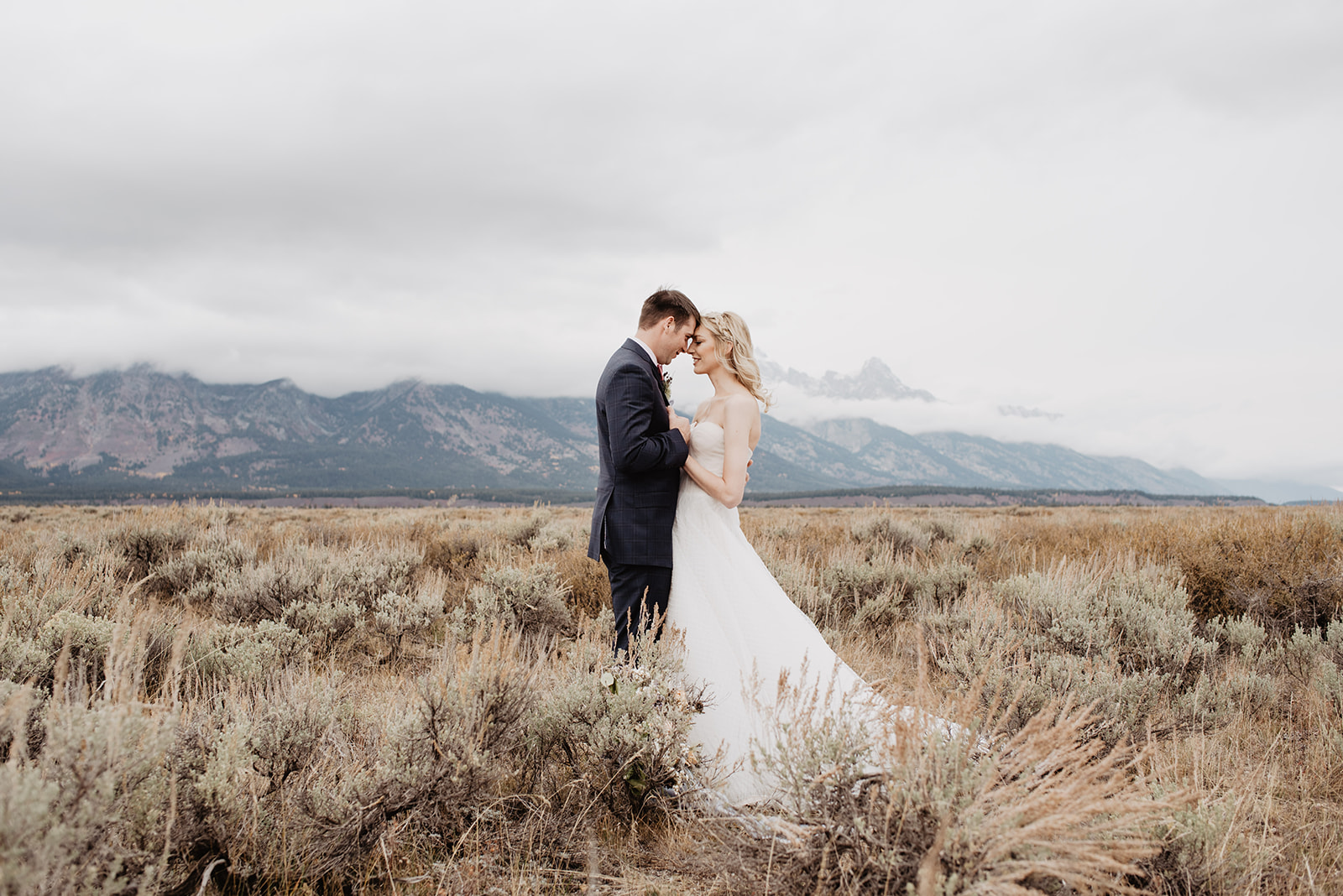 fall wedding day with bride and groom standing in a sage brush field in the Tetons while holding each other romantically with their heads together and the clouds from a storm drifting behind them