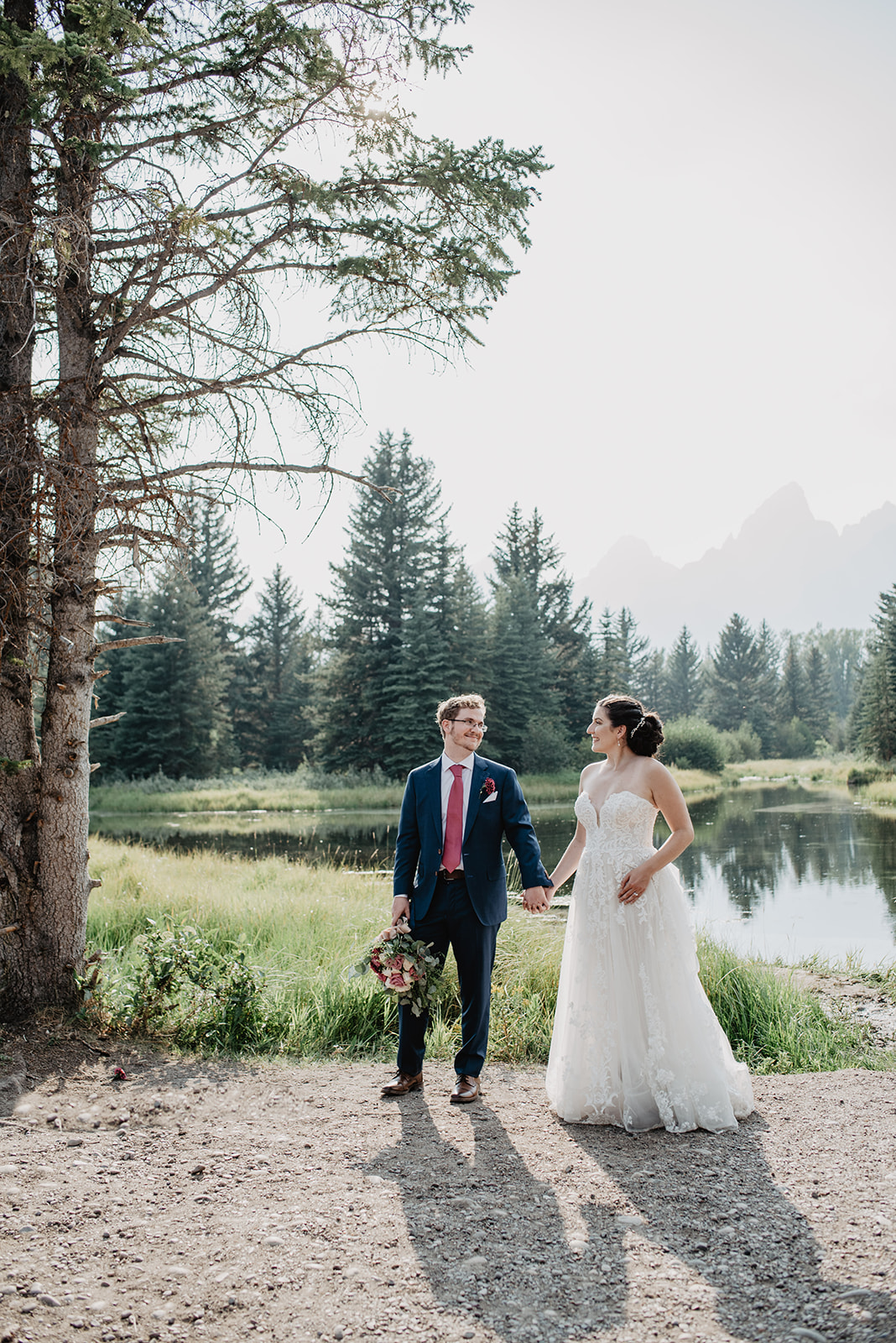 bride and groom holding hands together as they walk next to a lake in the Grand Tetons as they look at one another romantically