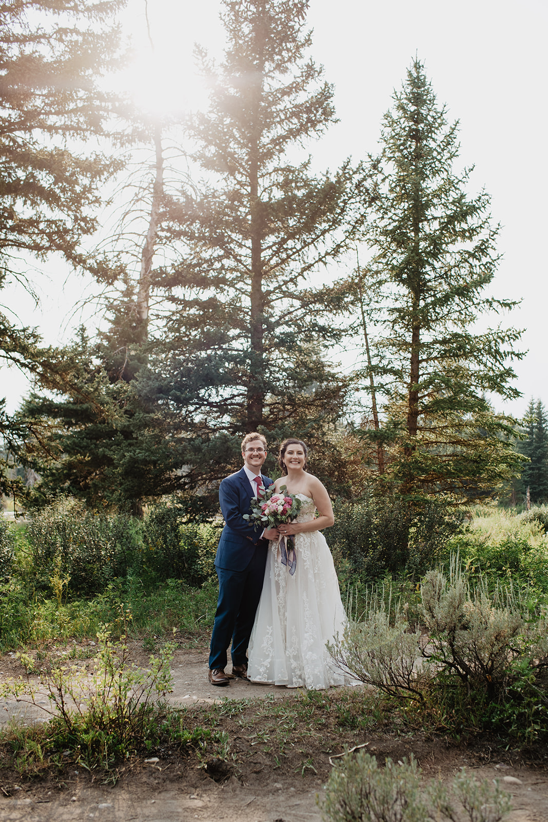 adventurous couple in the forest of the Grand Tetons on their elopement day standning side by side and smiling as the bride holds her florals and the sun shines behind them