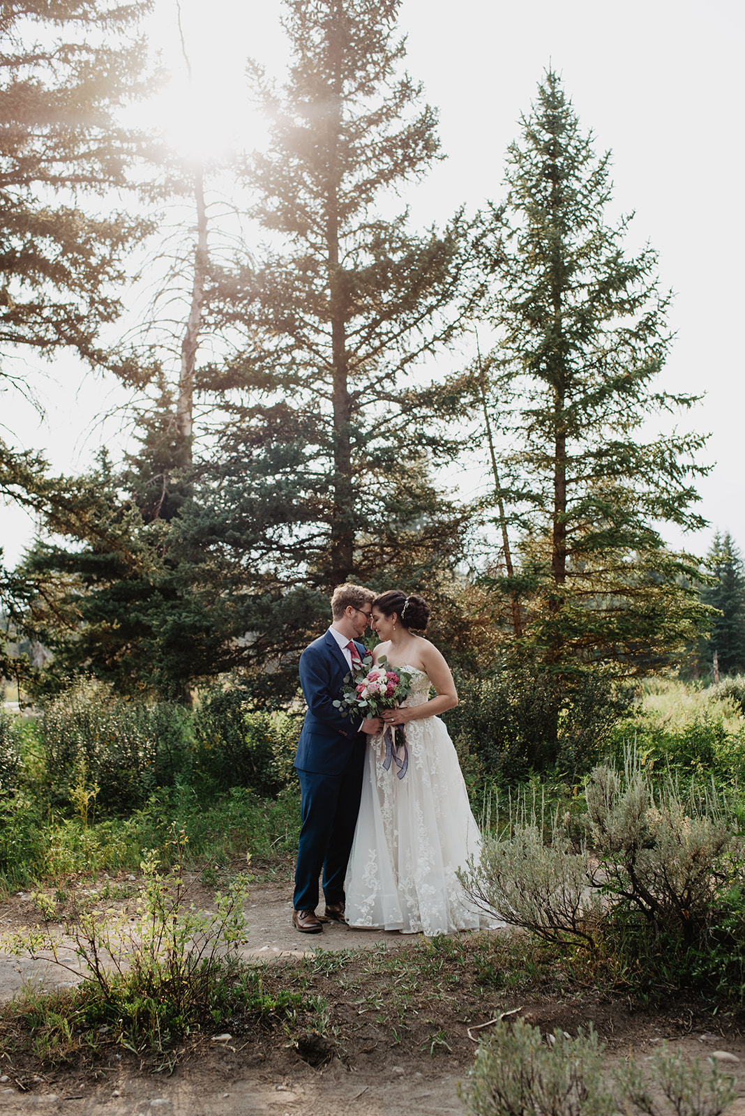 bridal portraits of man and woman embracing each other as they stand in front of some tall pine trees in Jackson Hole Wyoming with their foreheads touching as the bride holds her wedding bouquet