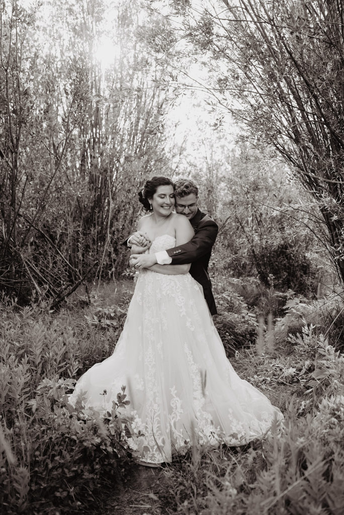 black and white image of bridal portraits in Schwabacher Landing in Jackson Hole Wyoming with groom hugging his bride from behind her and the bride smiling big as she looks back at her husband