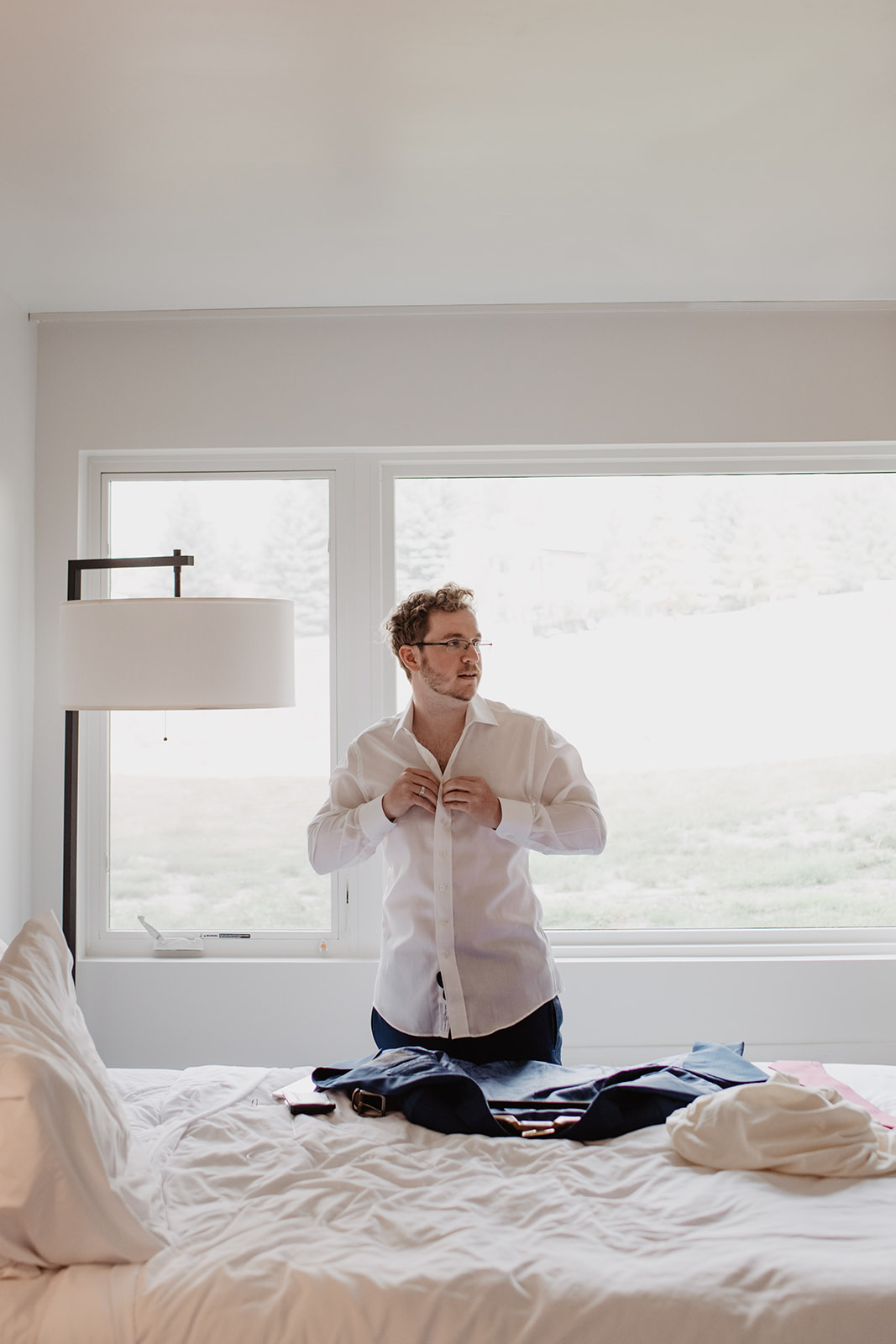 groom getting ready in his hotel room while buttoning his shirt and looking off to the distance in front of a large bright window