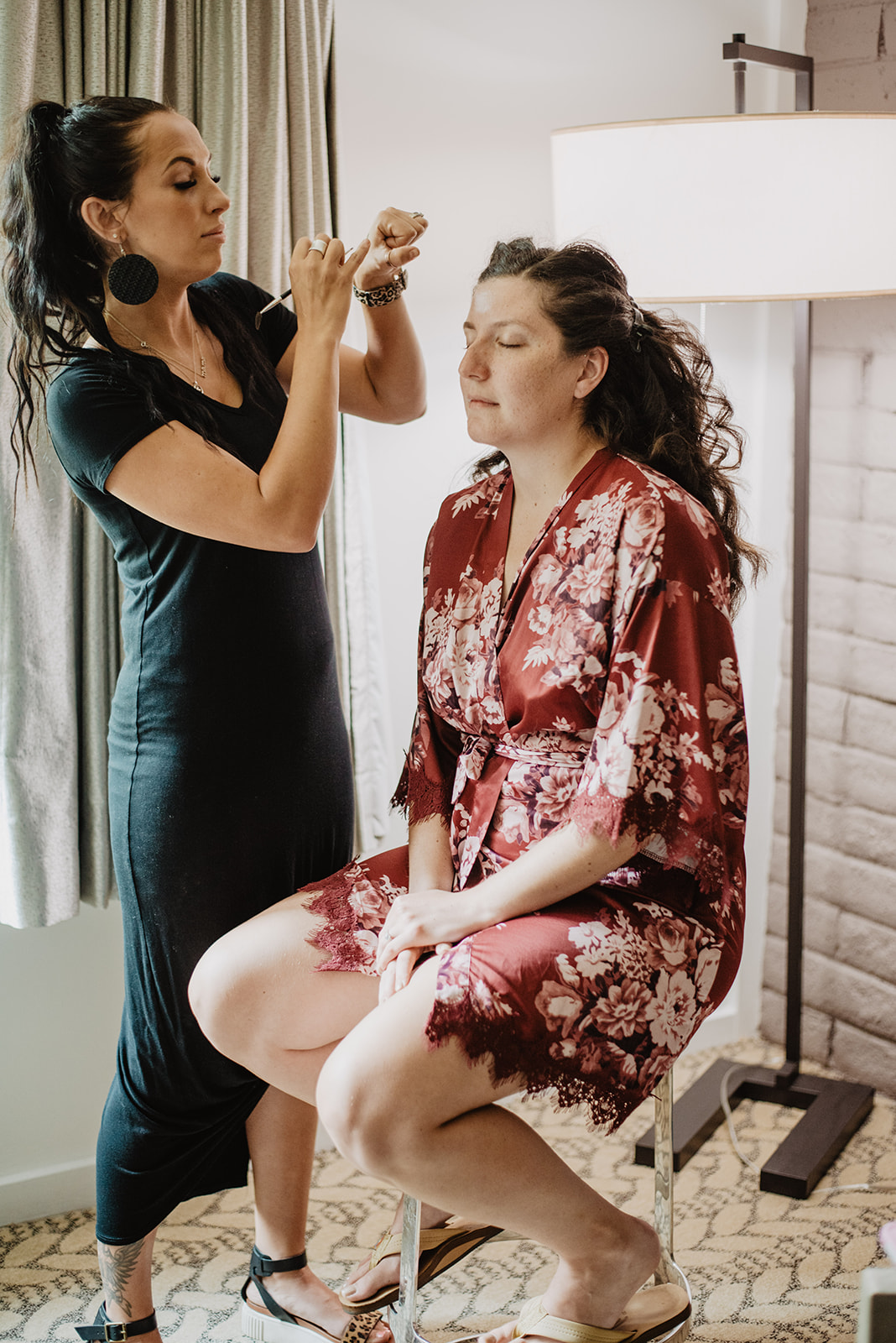 bride sitting on a stool while her make up artist from Jackson Hole does her makup in front of a window in her bridal suite