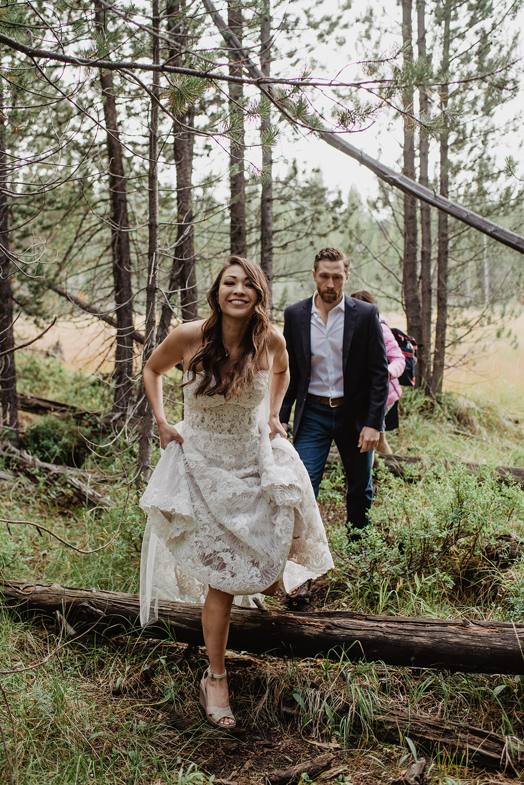 bride stepping over a fallen tree while hiking in her wedding dress in the Grand Tetons for her adventure wedding at Taggart Lake with her groom following behind her