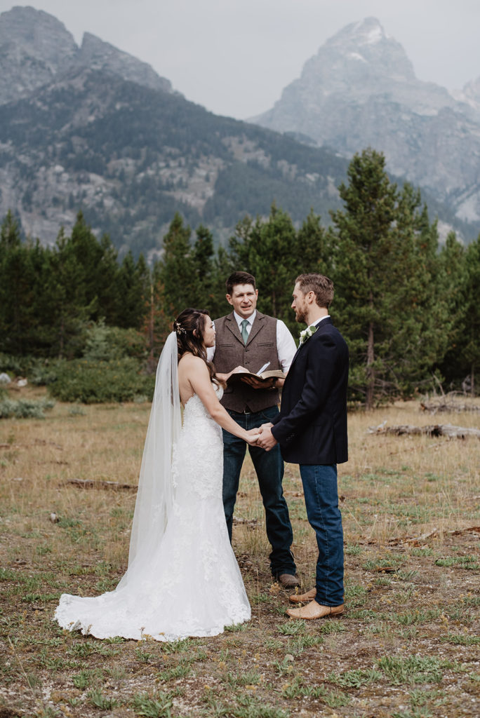 jackson hole photographers captures bride adn groom holding hands during their outdoor ceremony for their Grand Teton wedding  with the forest in the distance and the mountains towering over 