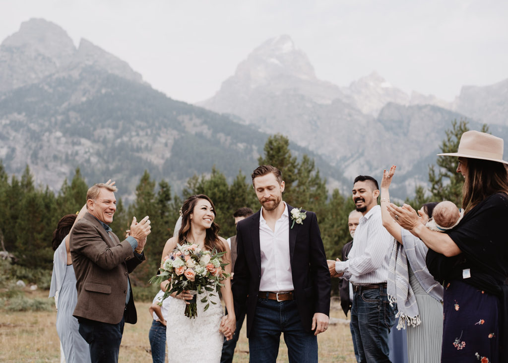 jackson hole photographers captures bride and groom holding hands and exiting their outdoor ceremony for their grand teton wedding in the woods with their family and friends cheering for them