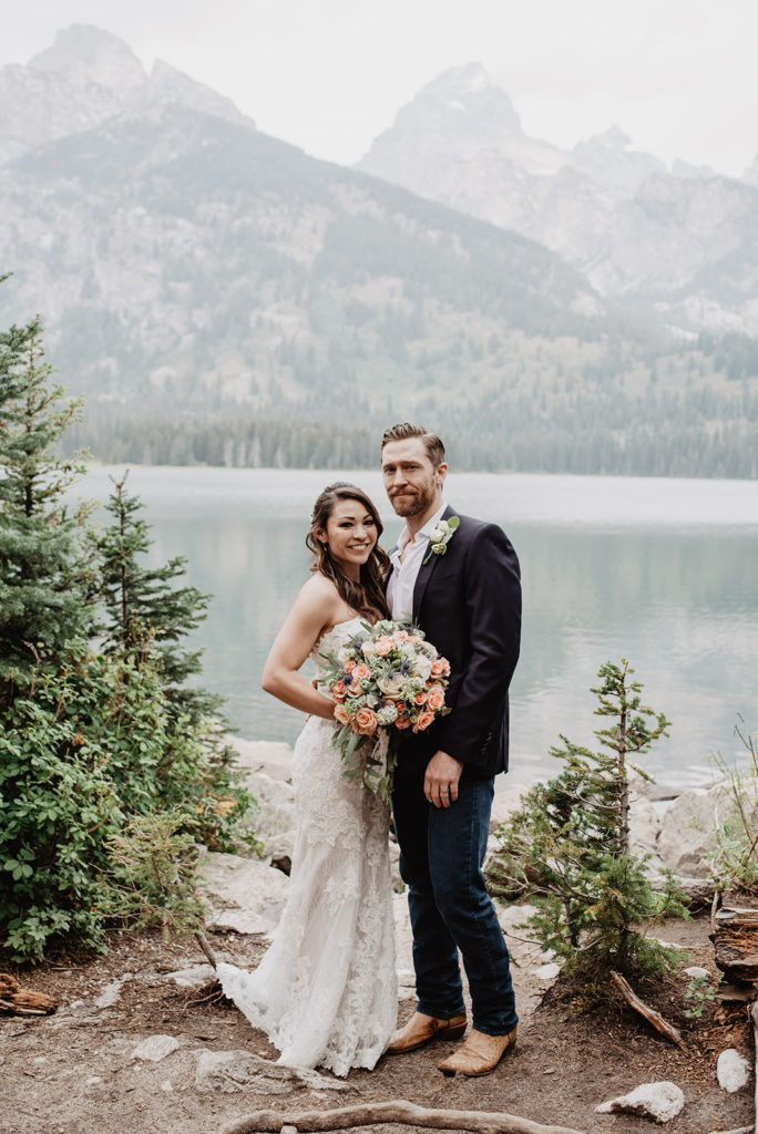 jackson hole photographers captures bridal portraits with bride and groom standing together at a lake in the Tetons and smiling as the bride holds a lush bouquet 
