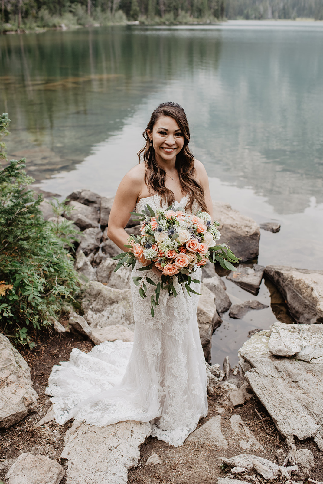 bride with a beautiful wedding bouquet with roses and thistles stands next to Taggart Lake in the Grand Tetons and smiles up at the camera