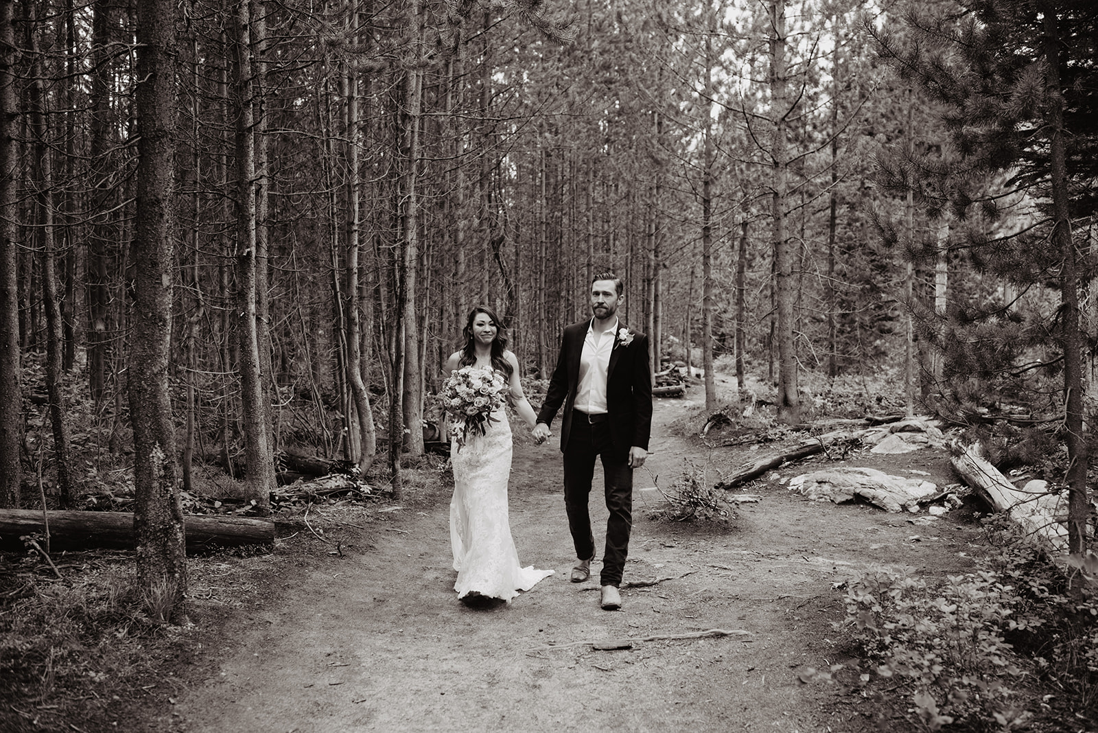 black and white image of bride and groom walking on a trail in the Grand Tetons with the forest behind them as they hold hands and smile in their wedding attire near Taggart Lake