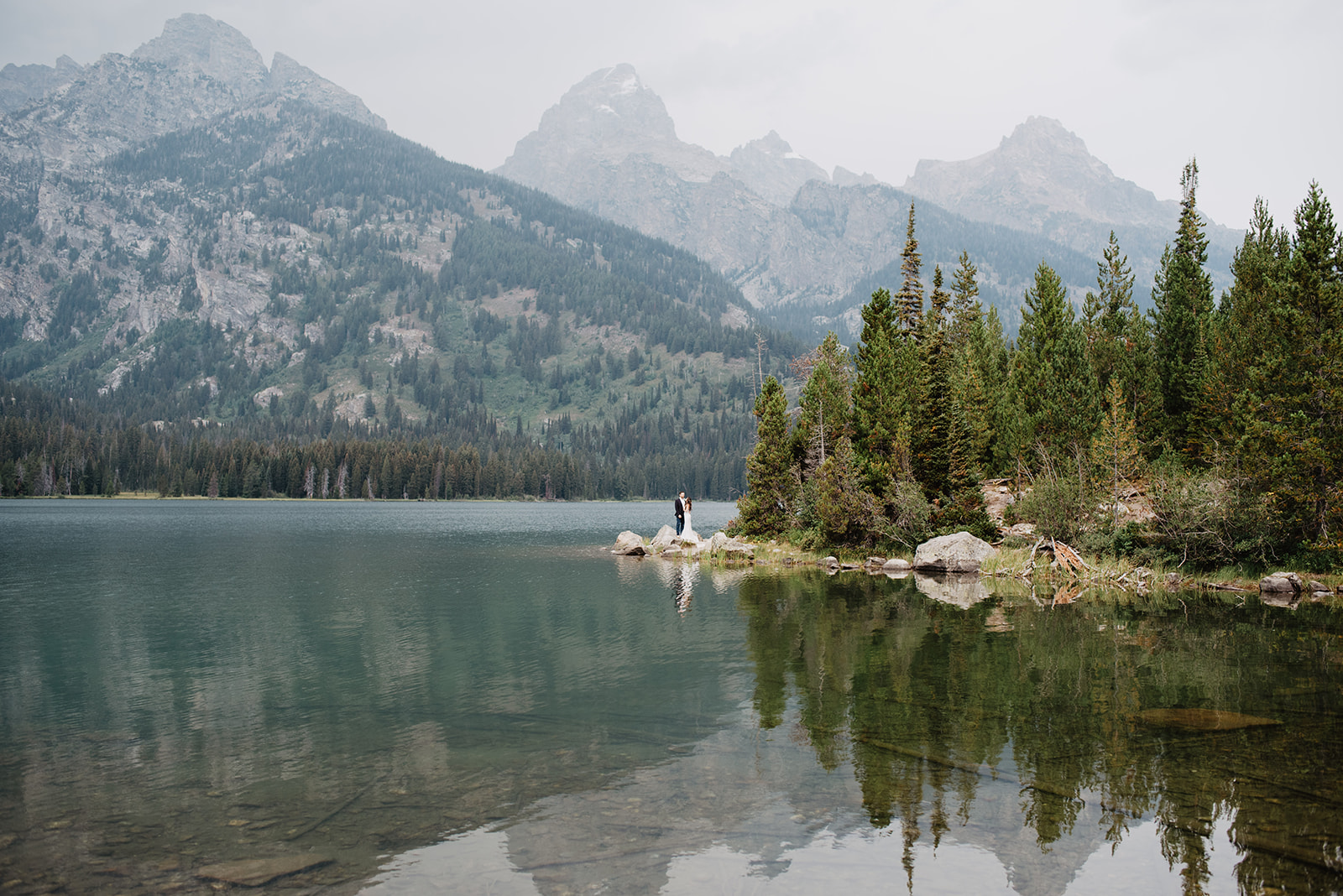 stunning image of Taggart Lake wedding taken by Grand Teton wedding photographer with bride and groom in the distance on a small rock in the lake and the Tetons reflecting into the water