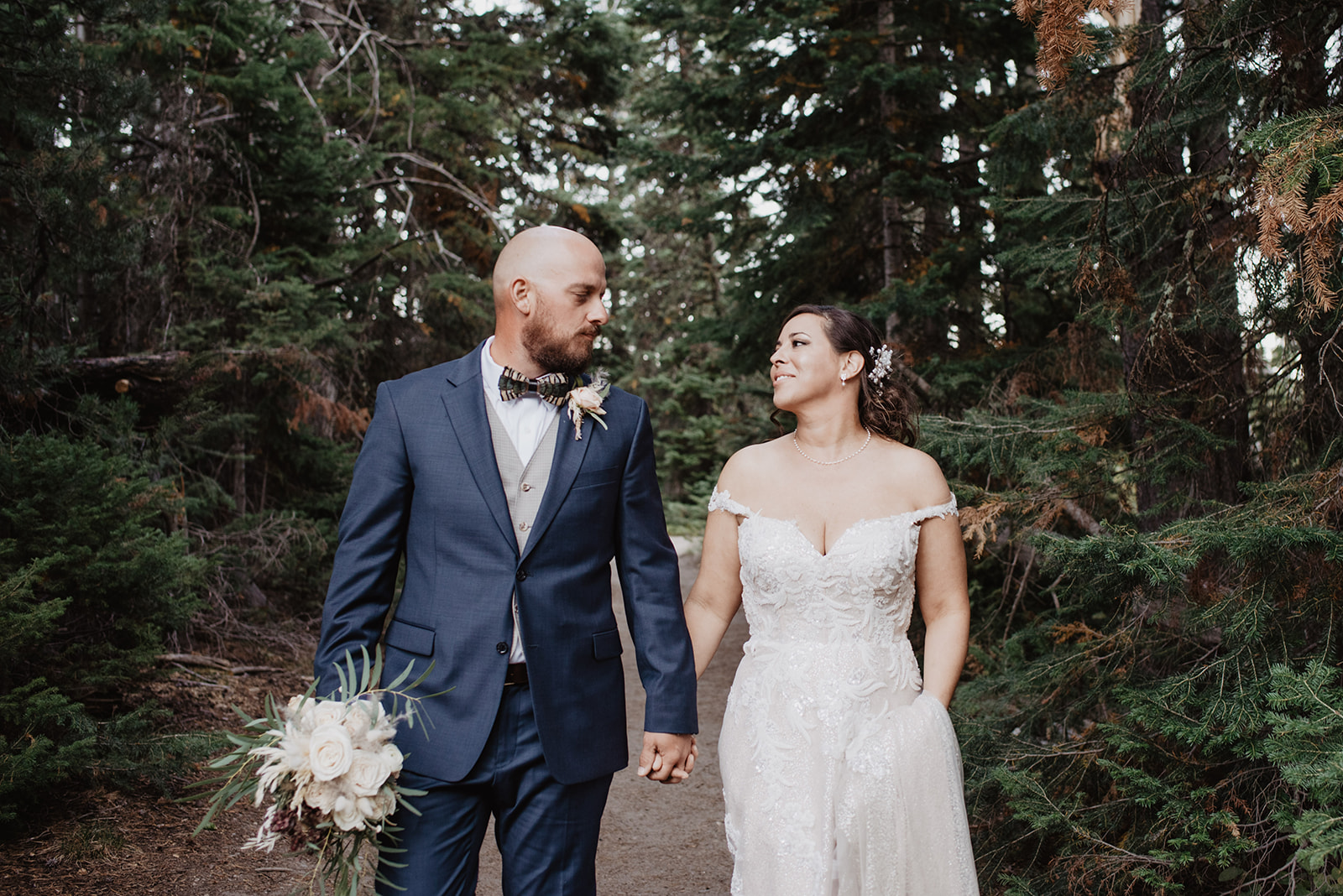 bride in a lace and sequence wedding gown holding hands with her groom who is in a blue suit as the groom holds the wedding bouquet and smiles at his bride for their JAckson Hole wedding while they walk in the woods 
