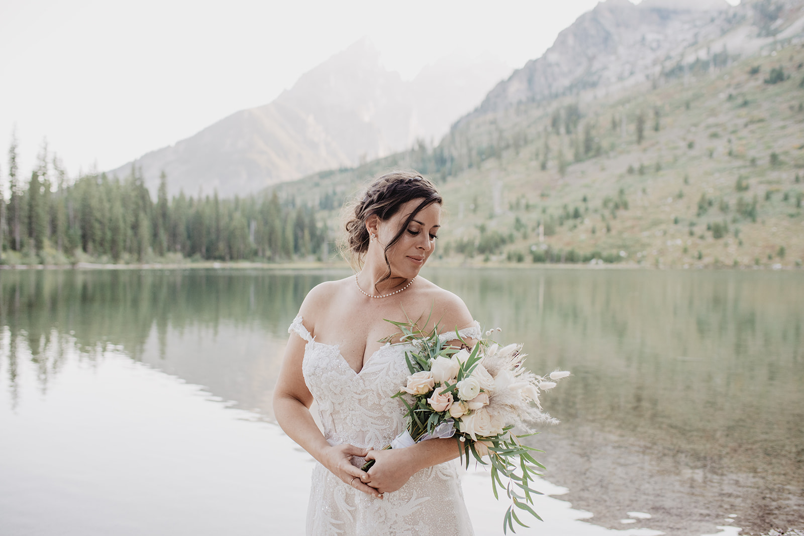 dark haired bride holding a wedding bouquet and looking over her shoulder at her flowers with the lake behind her with the Tetons reflecting in the water
