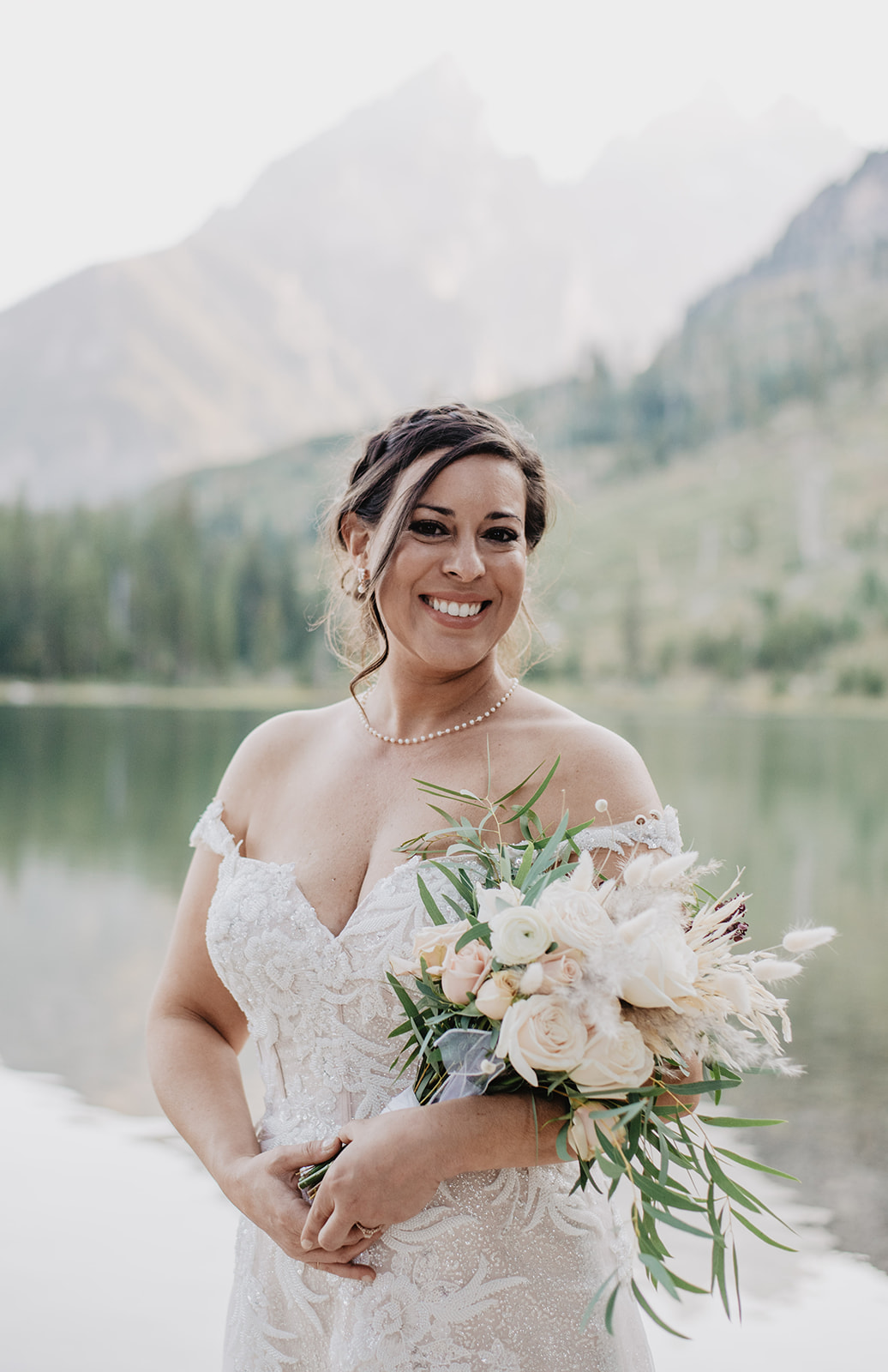 beautiful bride in lace wedding gown holding a bright white and pink wedding bouquet at String Lake for her bridal portraits with the Tetons and the water behind her 