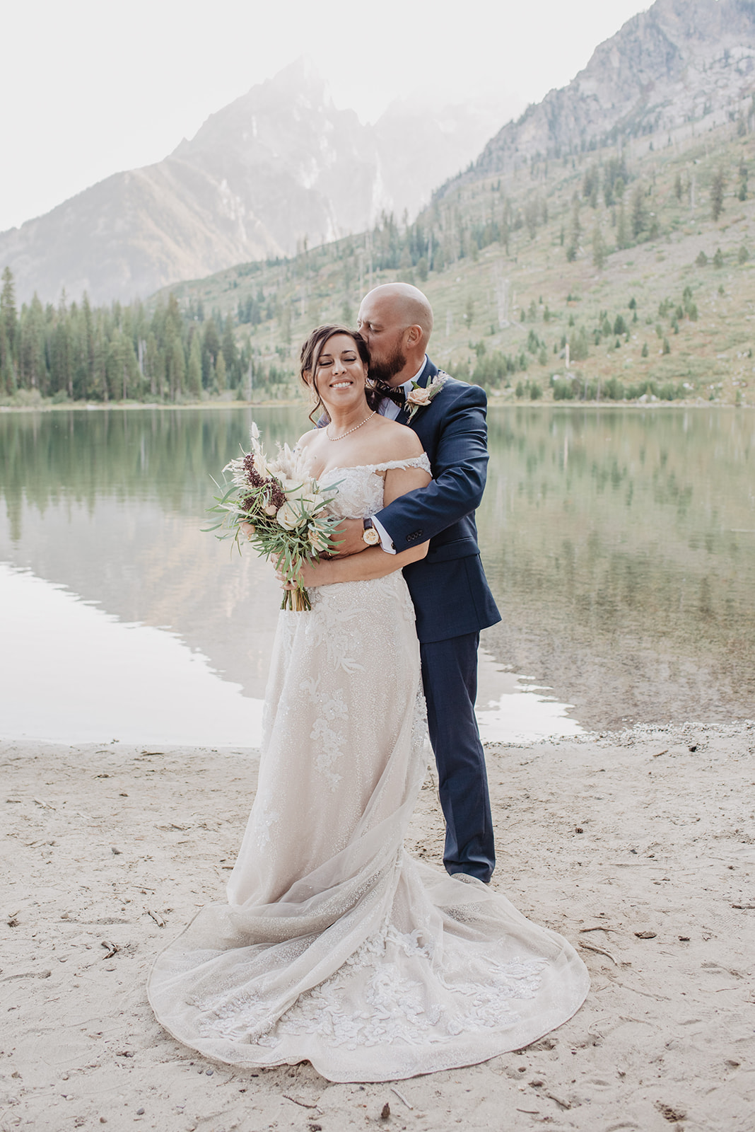 Oxbow Bend Elopement in the Tetons with bride and groom dressed up and standning next to String Lake as the groom holds onto the bride with the reflection of the mountain in the water
