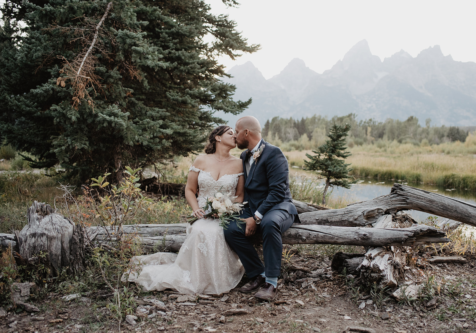 bride and groom sitting on a log bench in Jackson HOle for their Oxbow Bend Elopement in the Tetons as they kiss with the Grand Tetons mountains in the background 
