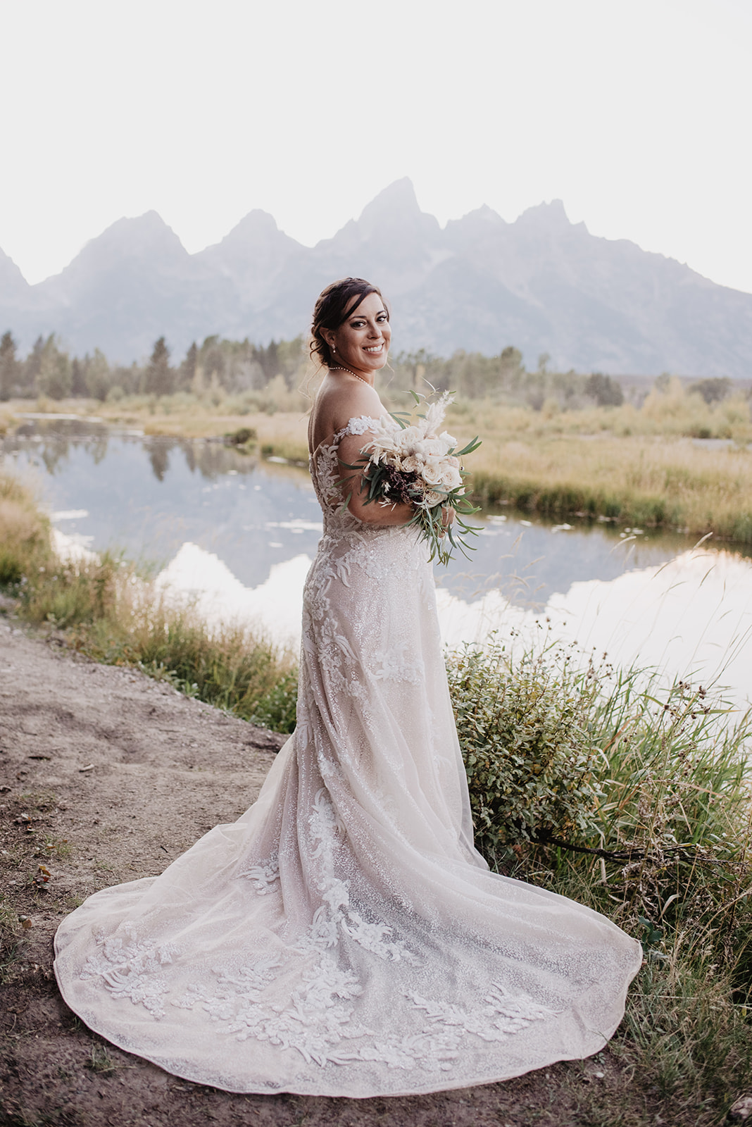 bridal inspiration with Jackson Hole bride standing next to a river and looking over her shoulder at the camera and smiling with her wedding florals as the Grand Tetons stand behind her