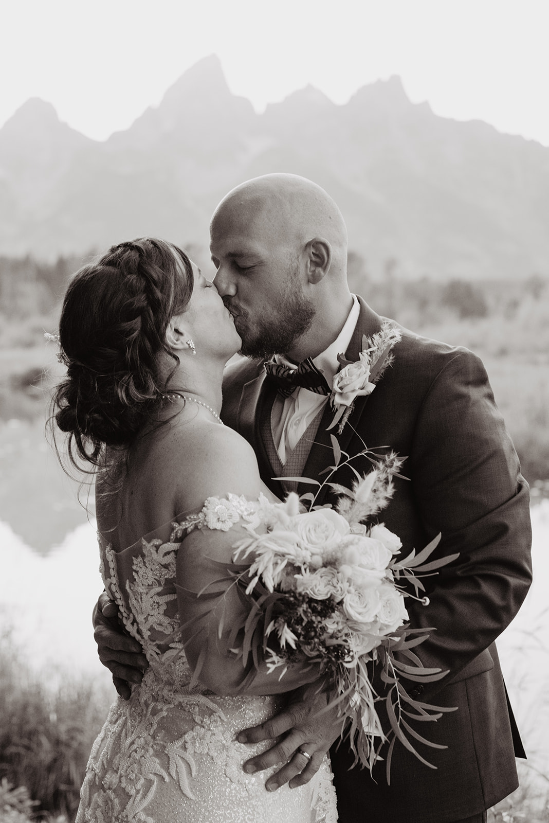 black and white wedding image of bride and groom kissing and embracing each other at Oxbow Bend for their wedding photos with the GR and Tetons behind them and reflecting in the water