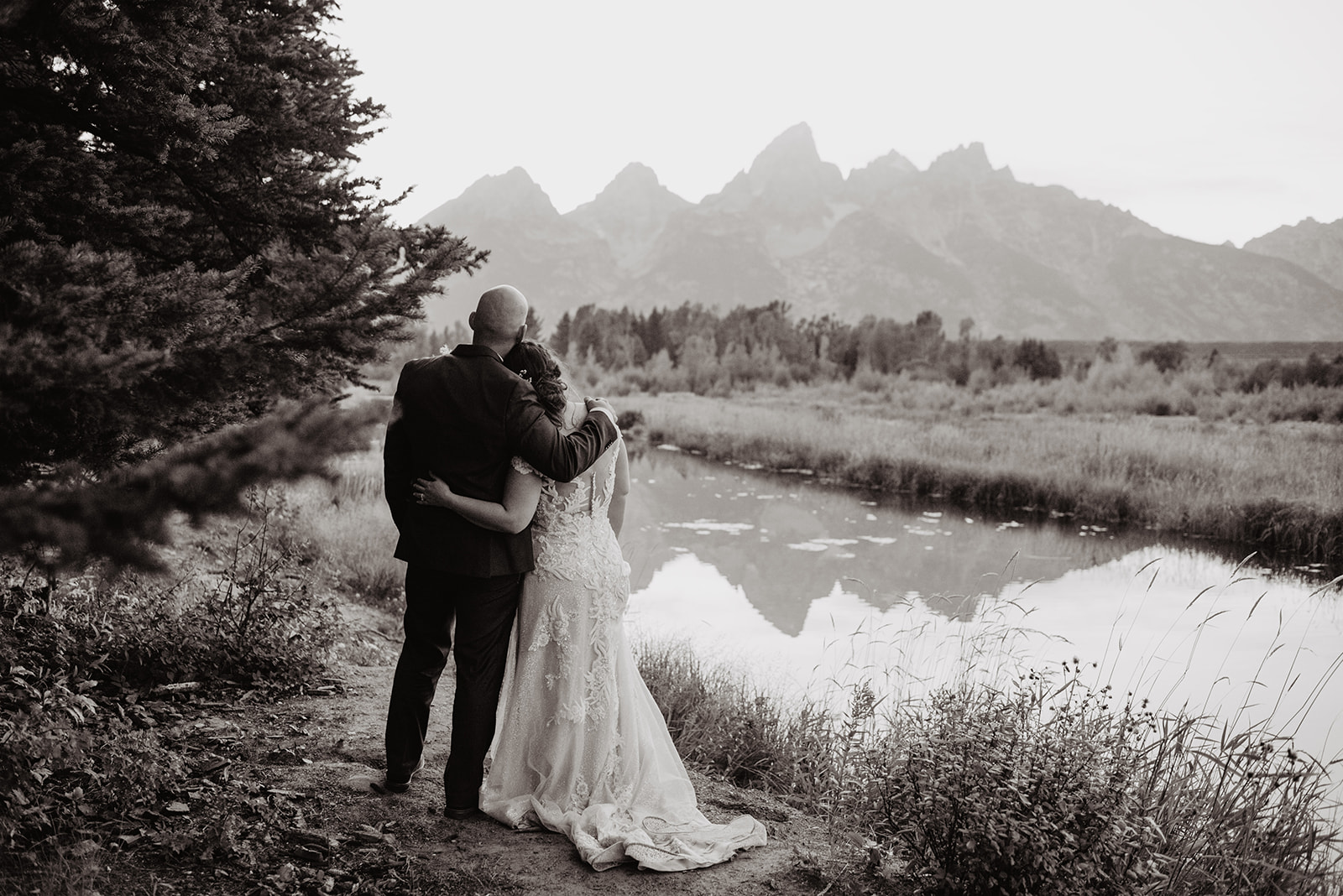 Grand Teton wedding day with bride and groom standing on the watter of a lake together and looking at the mountain range as they lean into eachother, captured by best Grand Tetons wedding photographers.