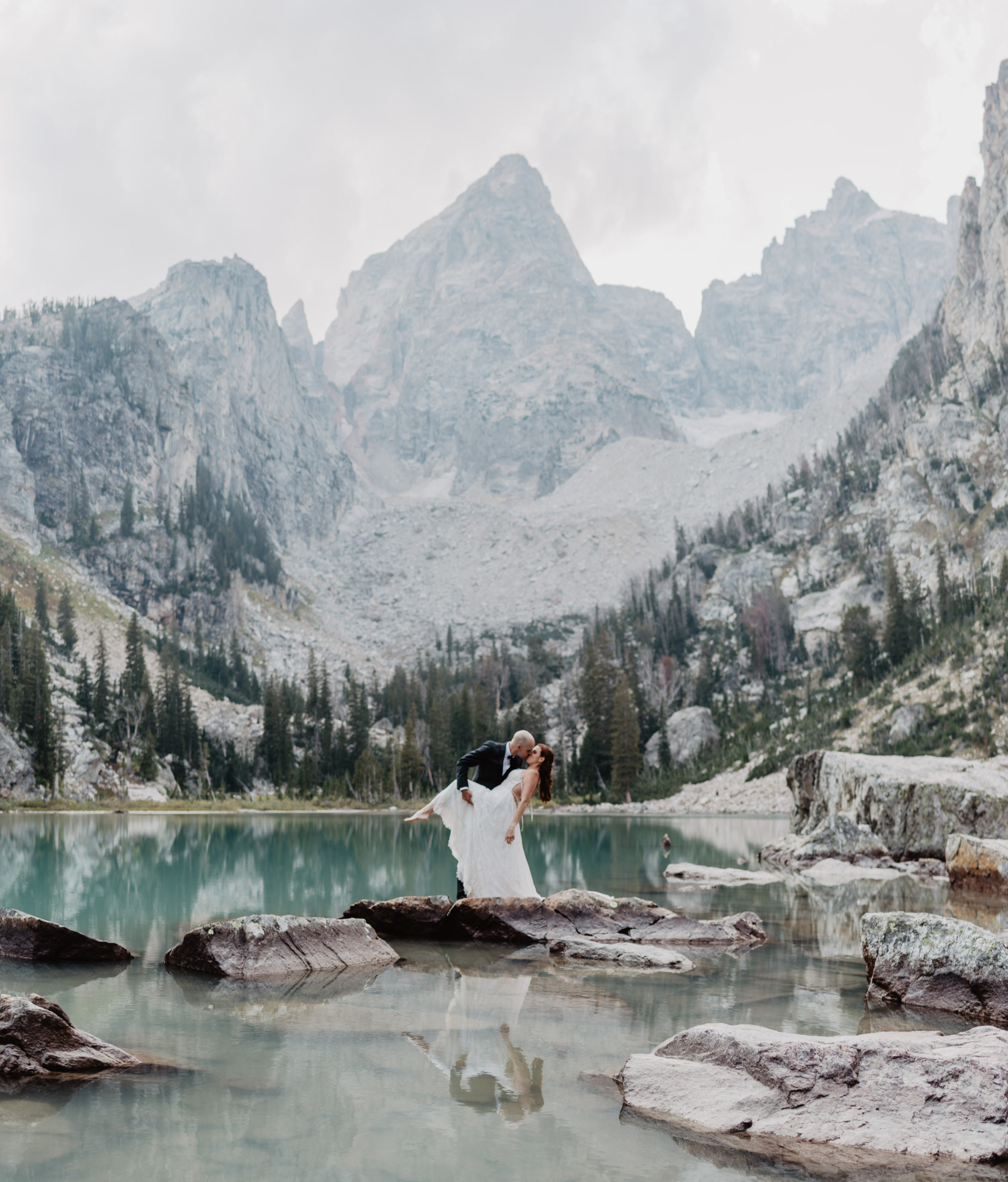 groom dips his bride and holds up her leg as they stand on a rock in the middle of Delta Lake with the Grand Tetons mountain range behind them