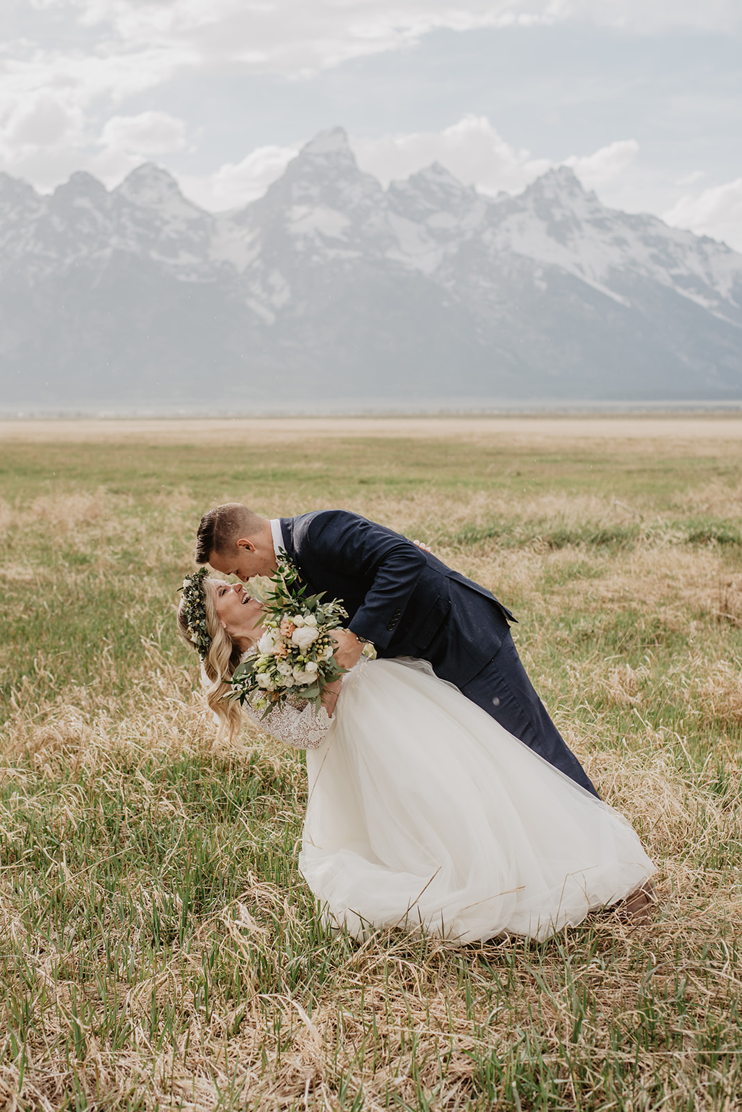 groom in a dark blue suit dips his bride as they laugh together in a meadow in the Grand Tetons for their summer wedding with bride in a 2022 wedding dresses