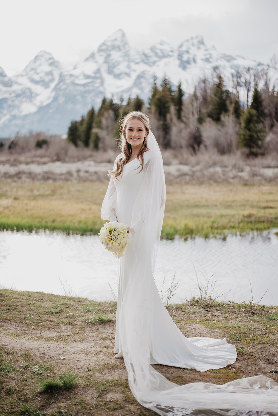 simple satin wedding dress on a beautiful bride in the Tetons as she stands next to a river on her wedding day captured by best JAckson Hole wedding photographer